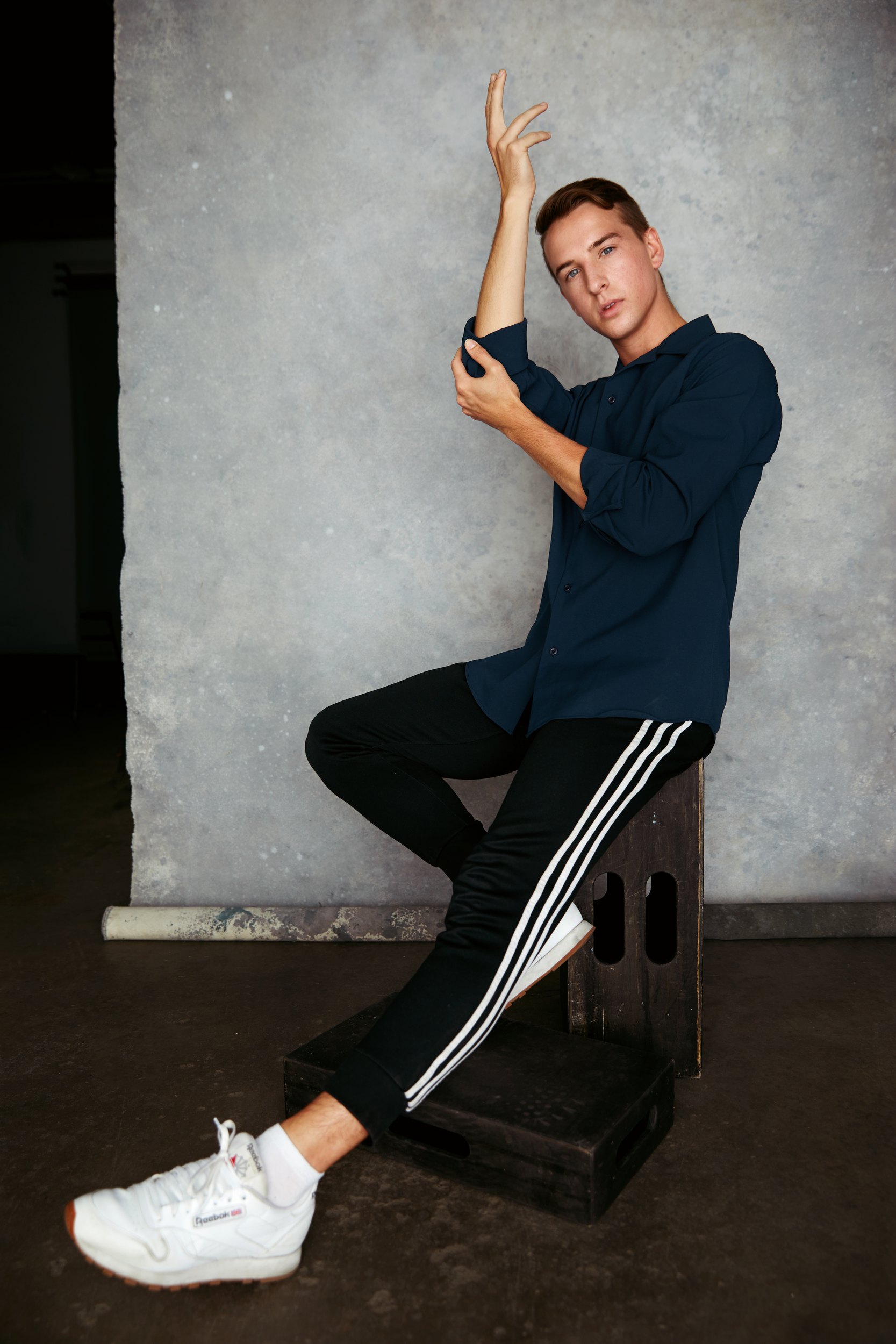 The navy shirt and track pants, seated on a black box against grey, one arm raised in a curve