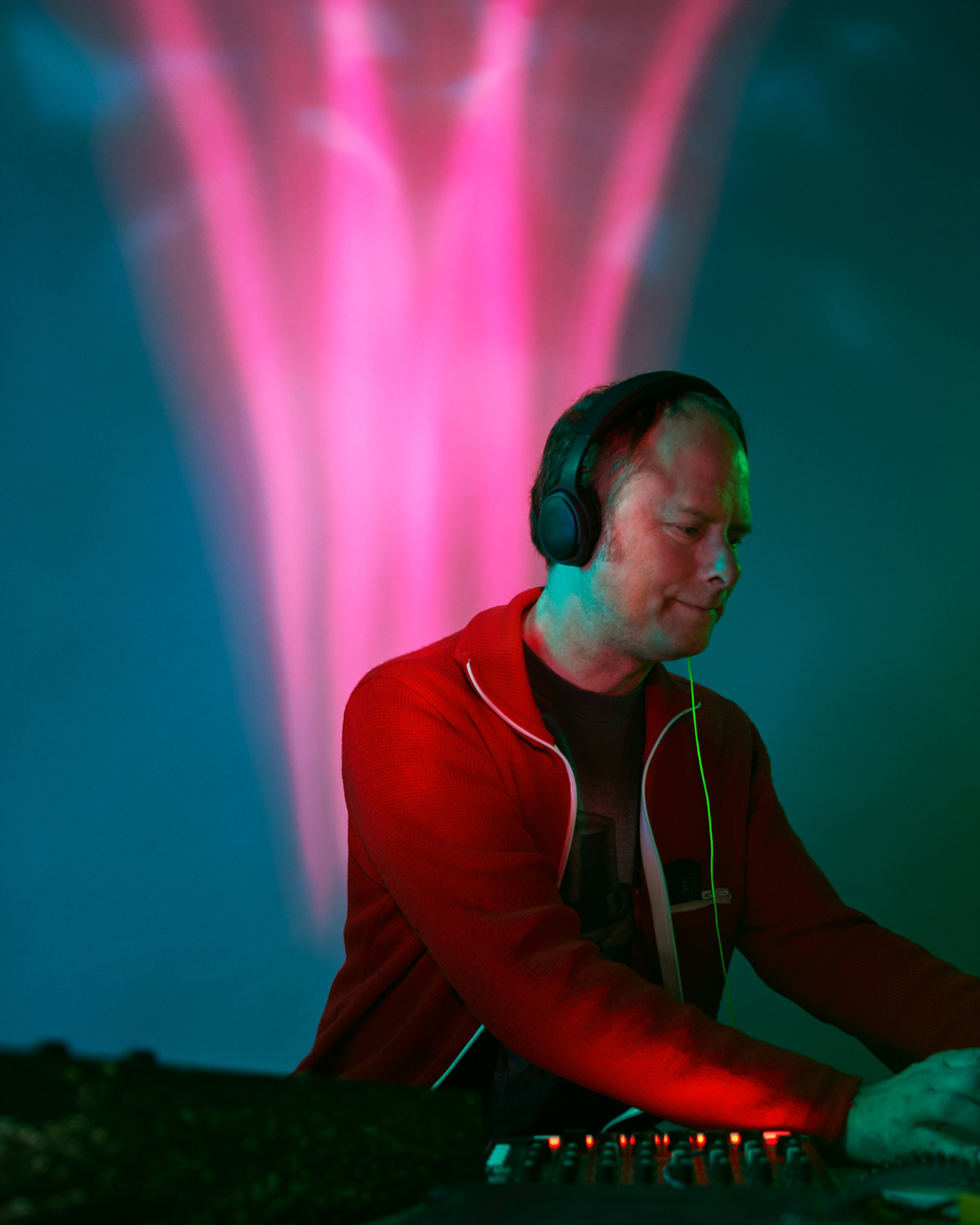 A musician in headphones and a red jacket at his equipment, a column of pink light behind him