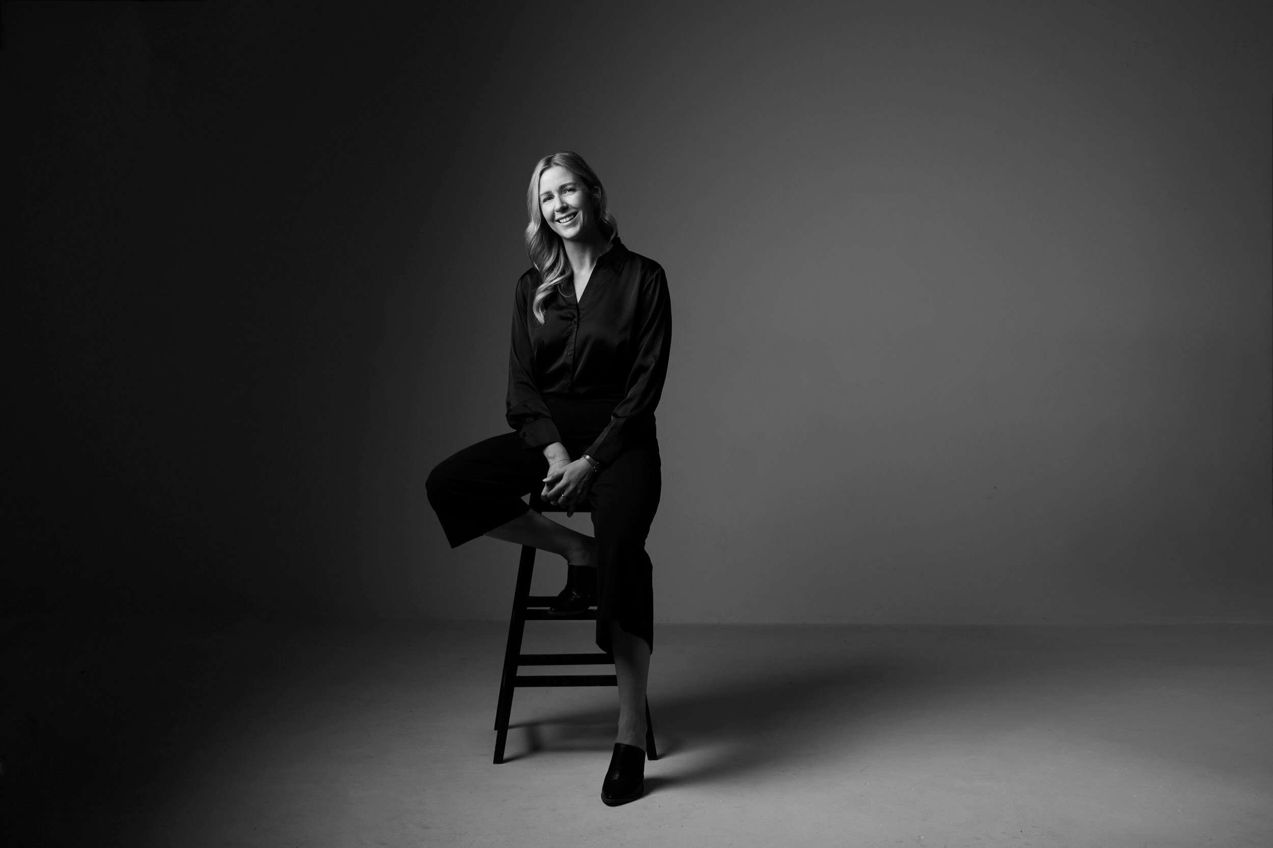 A portrait for BOMA, black and white: a woman seated on a stool in a dark dress, against grey