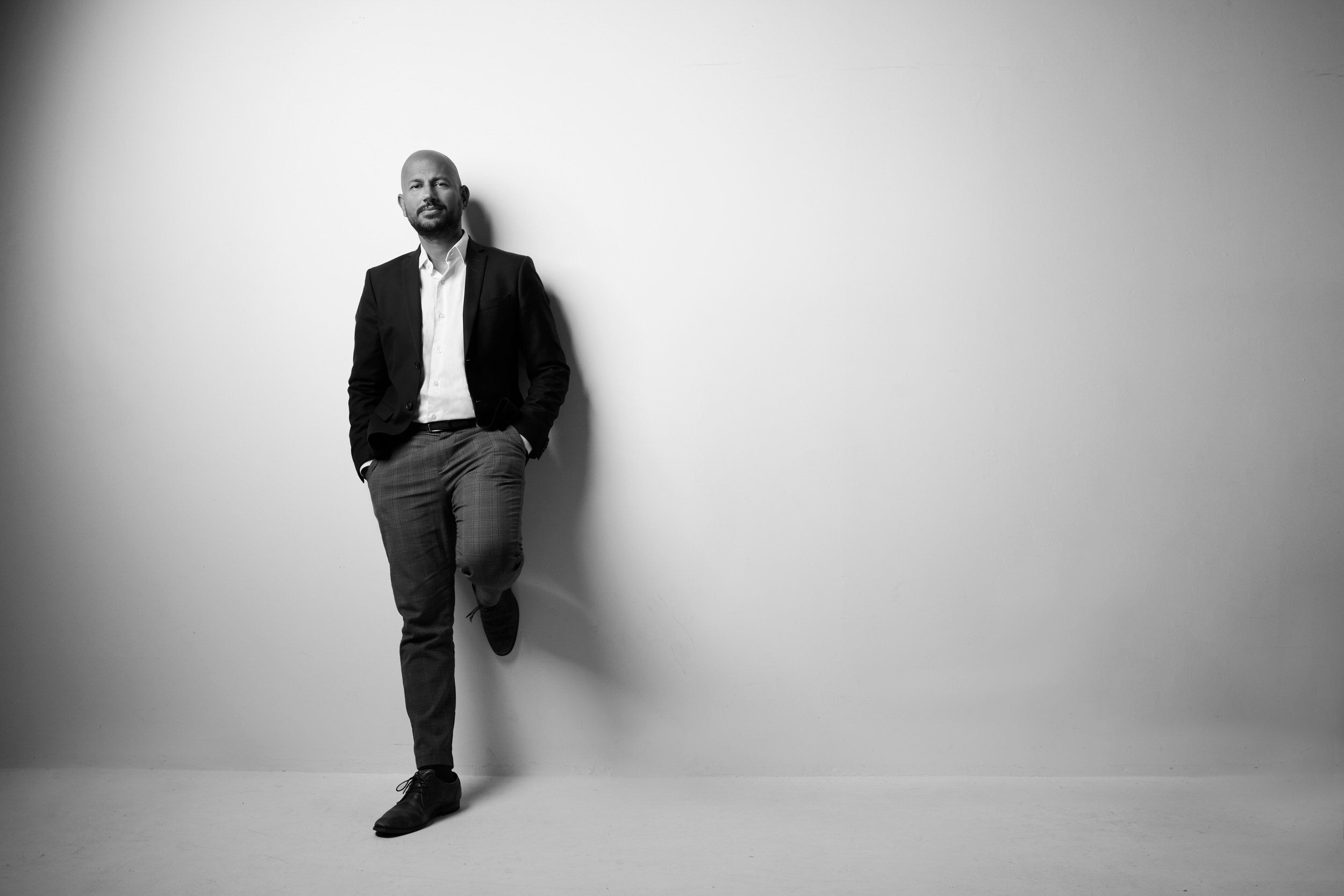 A portrait for BOMA, black and white: a man leaning on a white wall, hands in pockets, full length