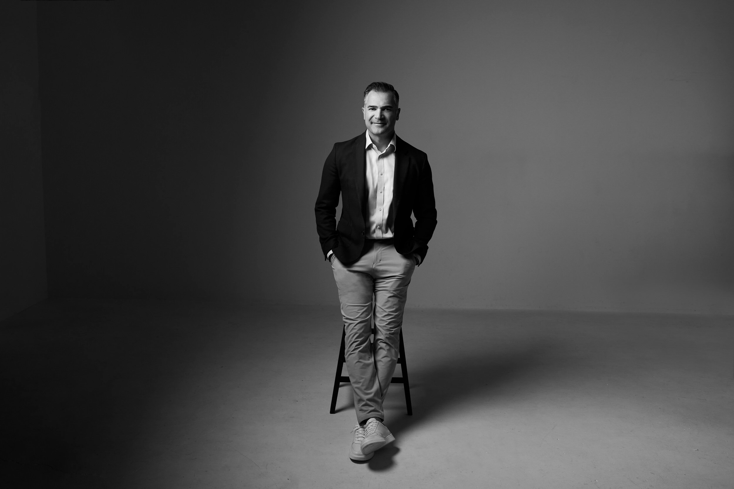 A portrait for BOMA, black and white: a man seated on a stool in a dark jacket, against grey