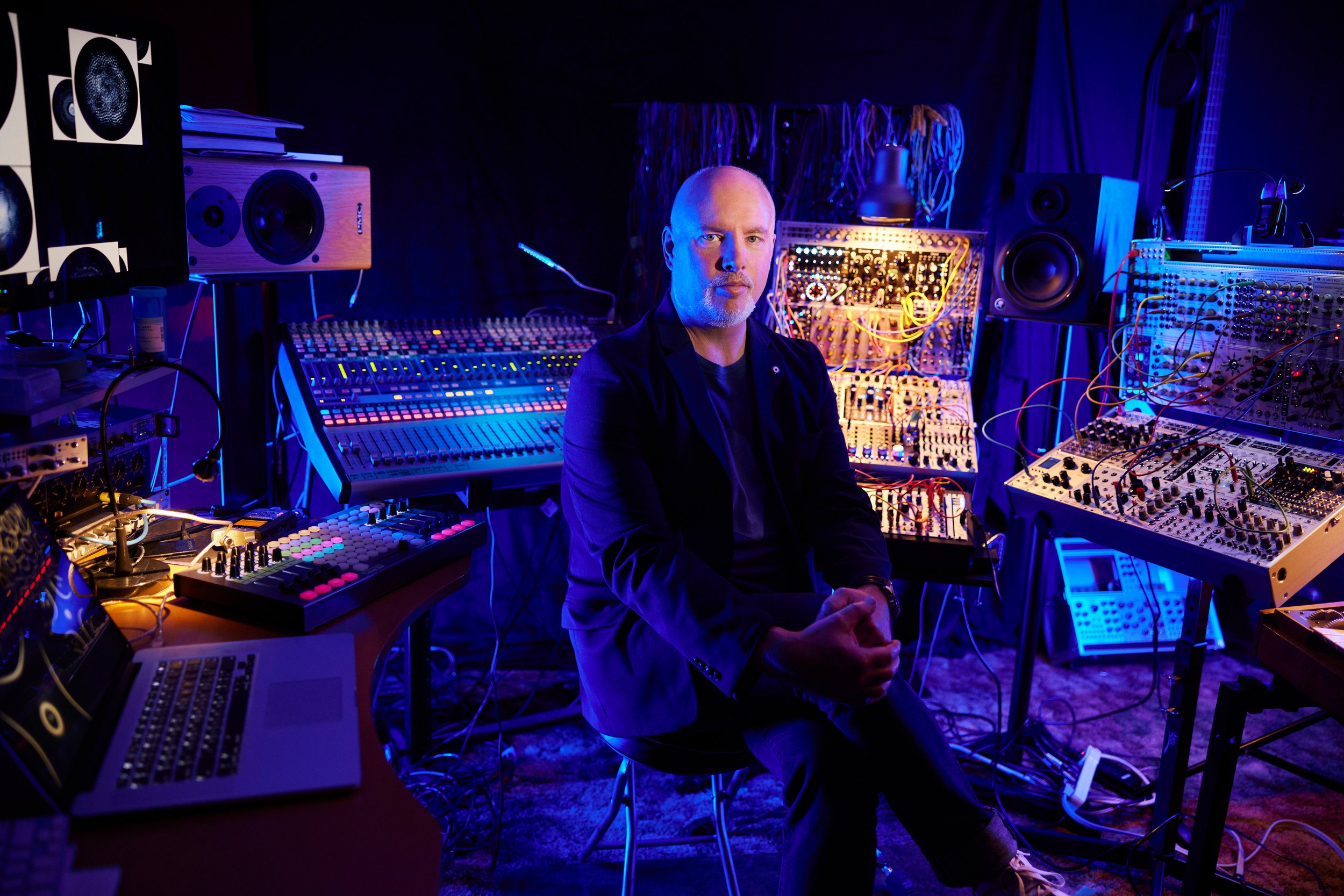 Gary James Joynes seated among his synthesizers and speakers, lit blue