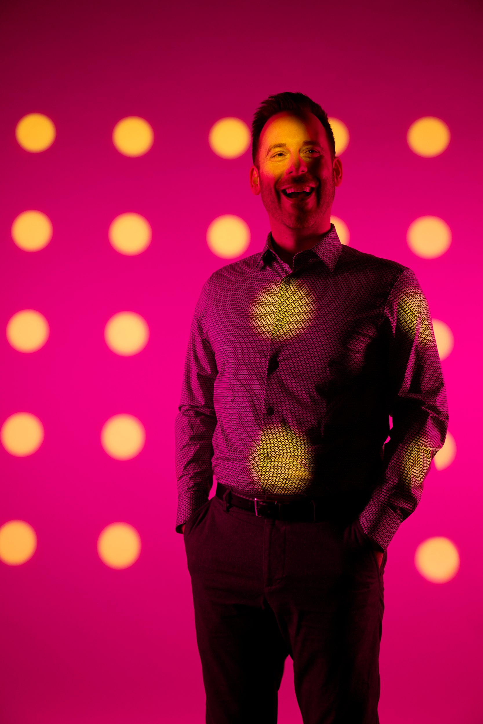 Standing, hands in pockets, lit through the card from the left
