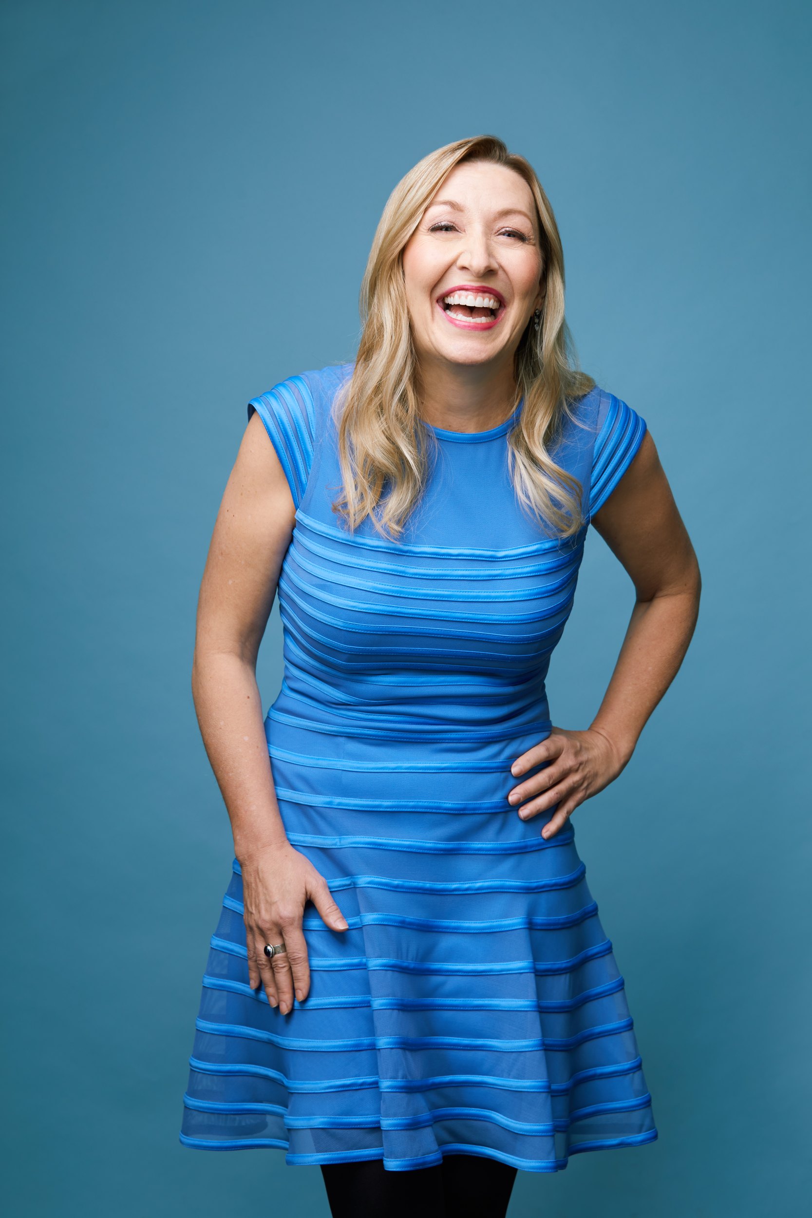 Dr. Jana Rieger laughing in a blue dress against blue, hands on her hips