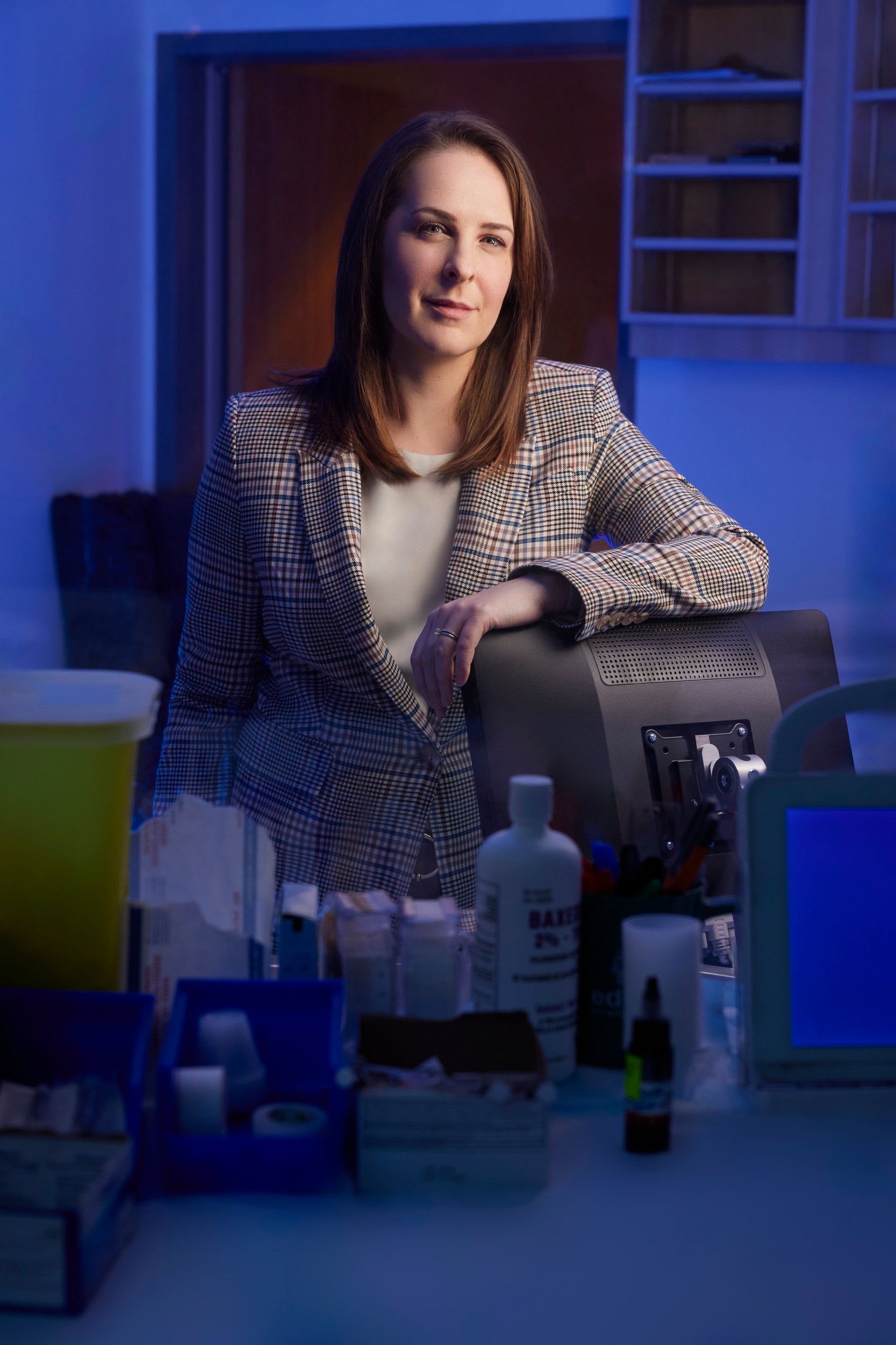 Seated at a lab bench, an arm along the back of the chair, bottles on the shelf behind