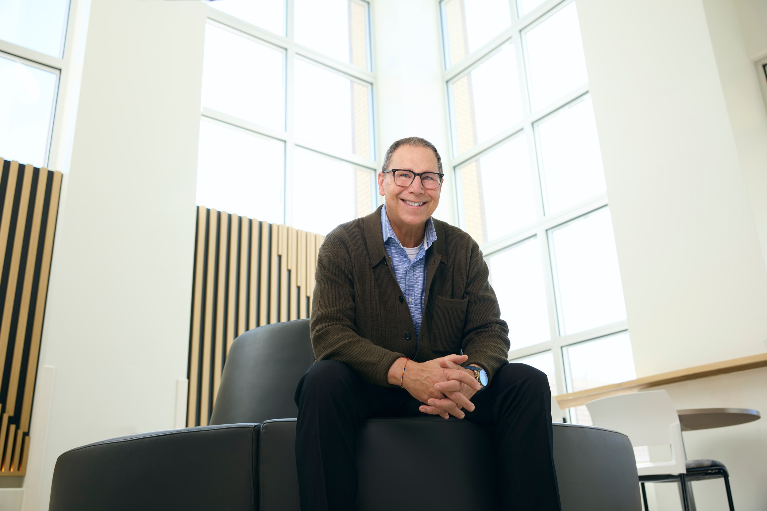 John Sexsmith seated in a bright atrium, hands folded, the tall windows behind him