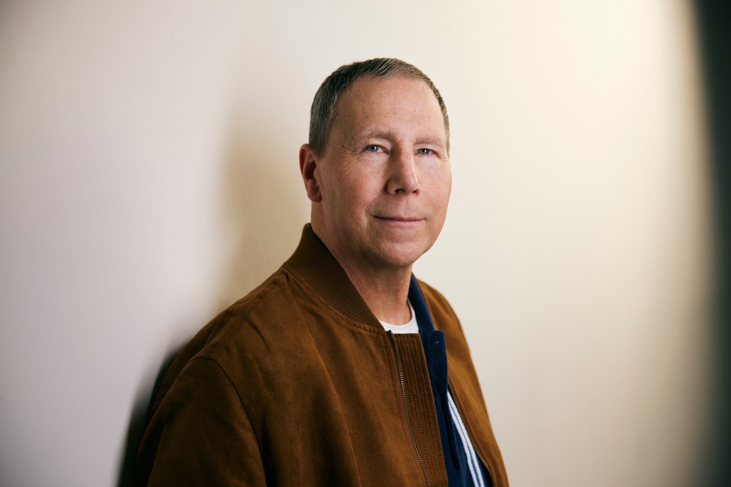 John Sexsmith in a brown suede jacket against a white wall, the light falling across him