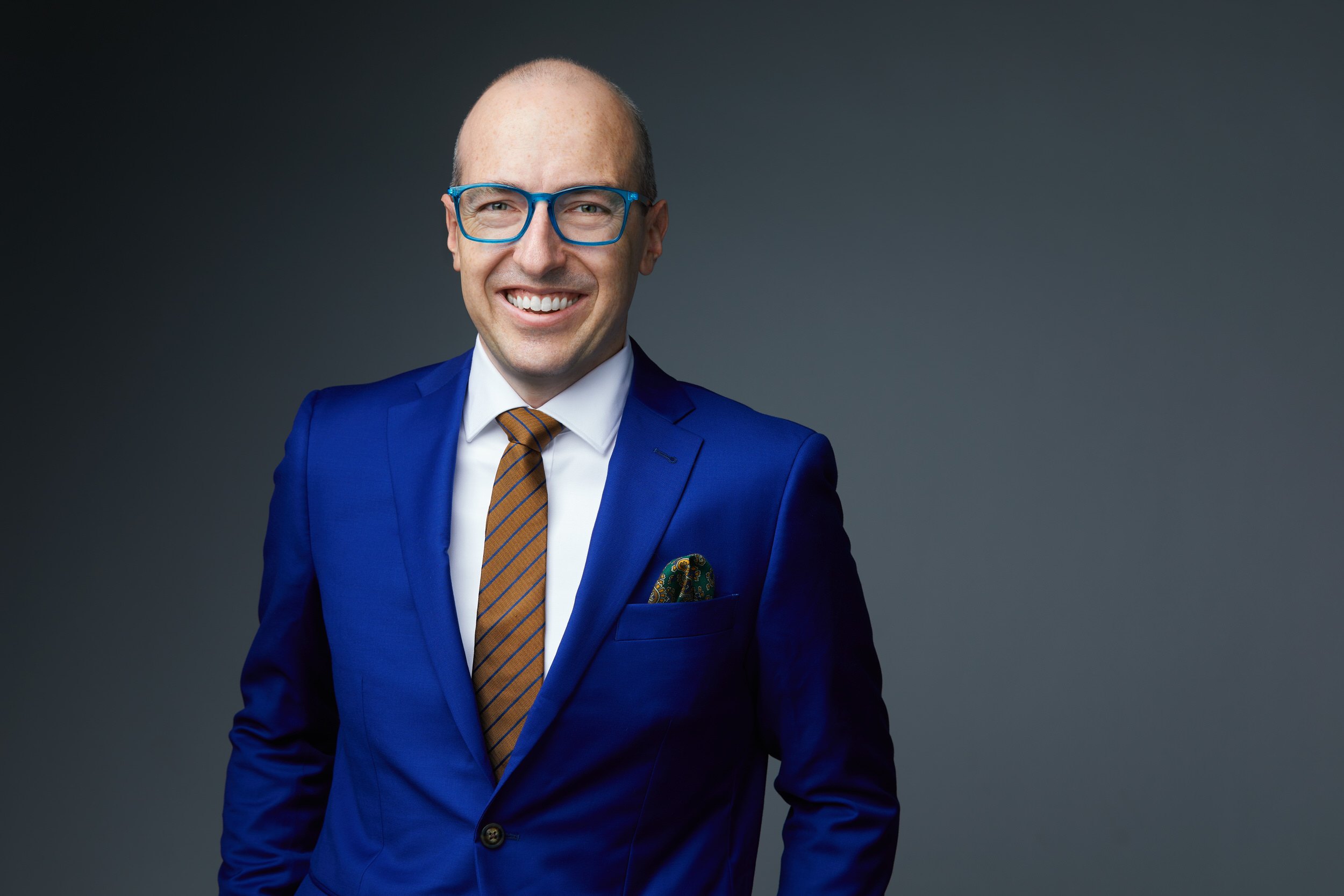 Leadership portrait: a blue suit and a patterned tie, standing, smiling, against charcoal
