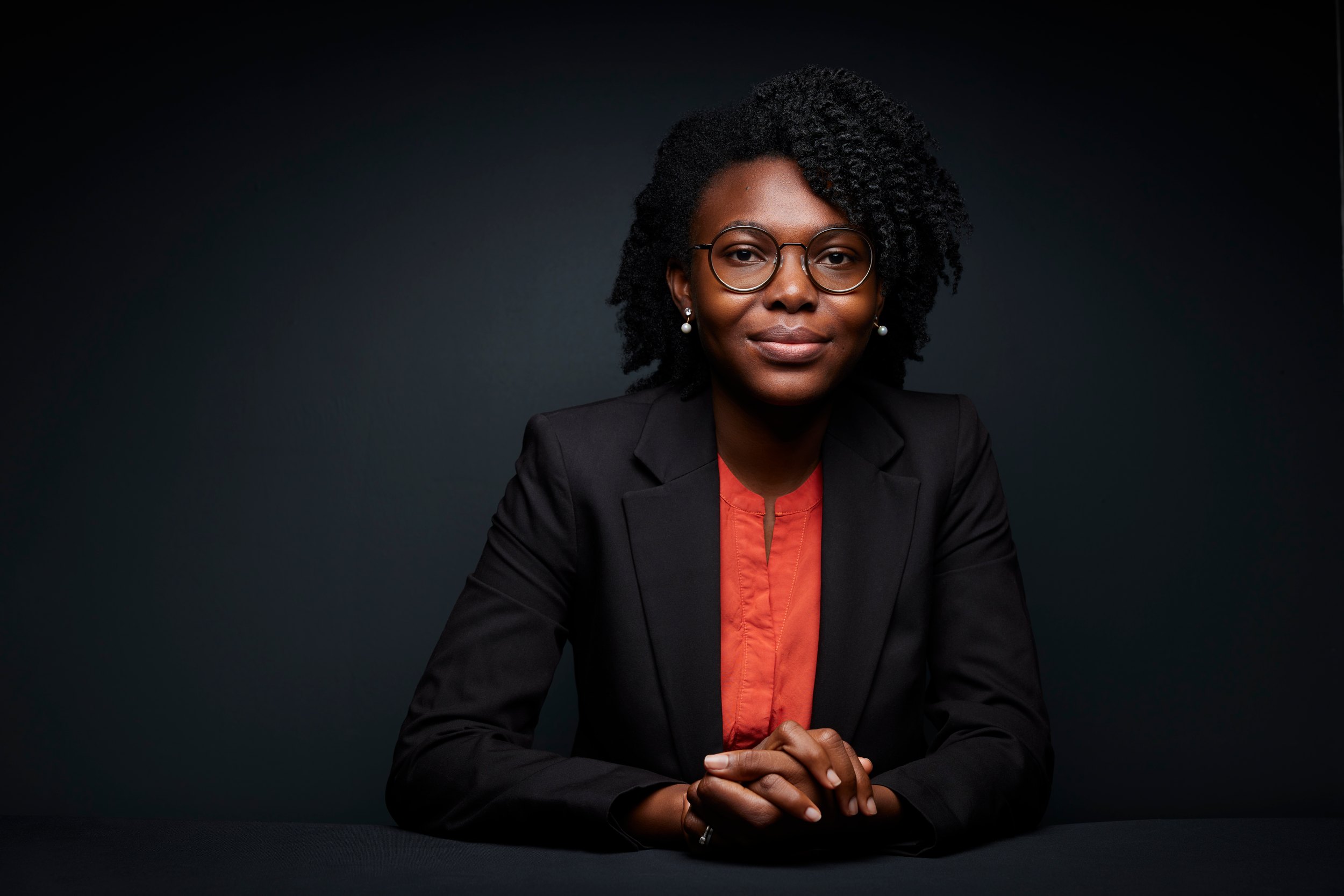 Leadership portrait: seated, a black blazer and an orange shirt, low light