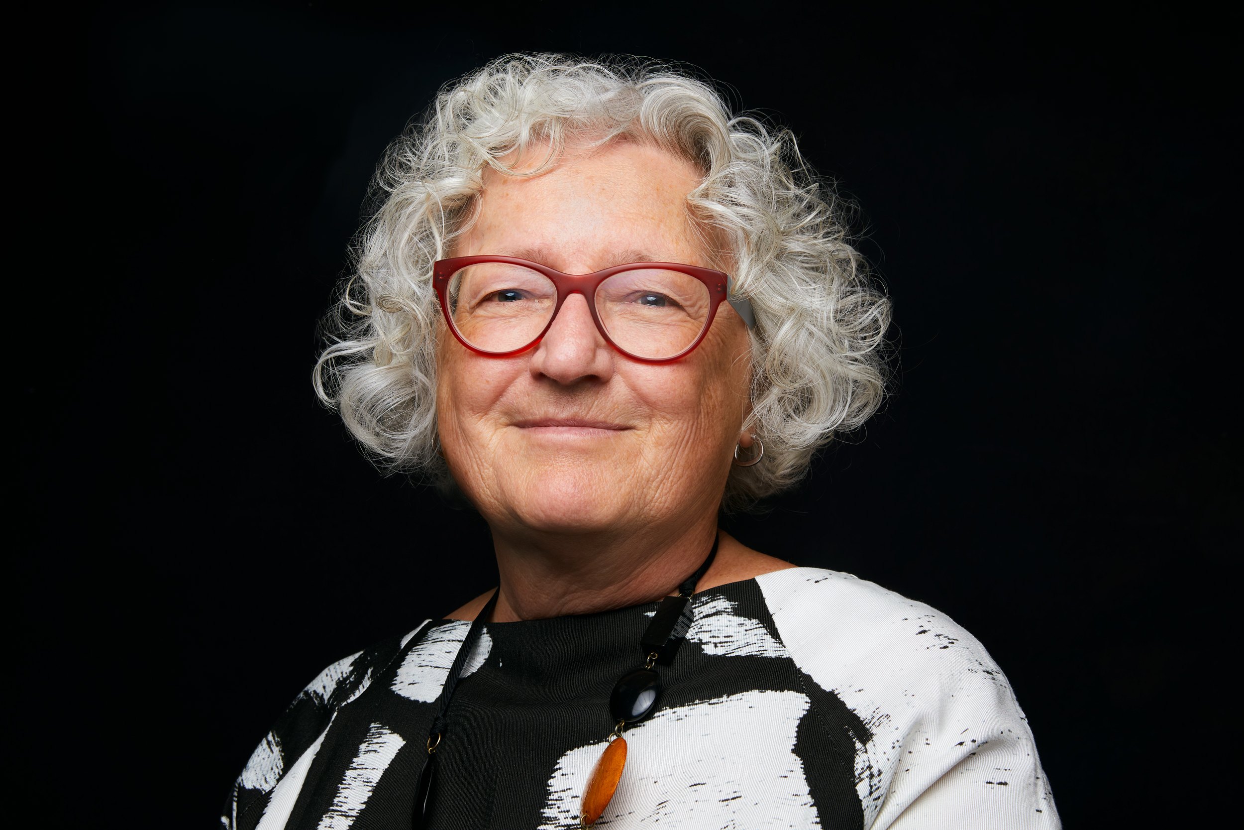 Leadership portrait: grey curls, red glasses, a black and white top, low light