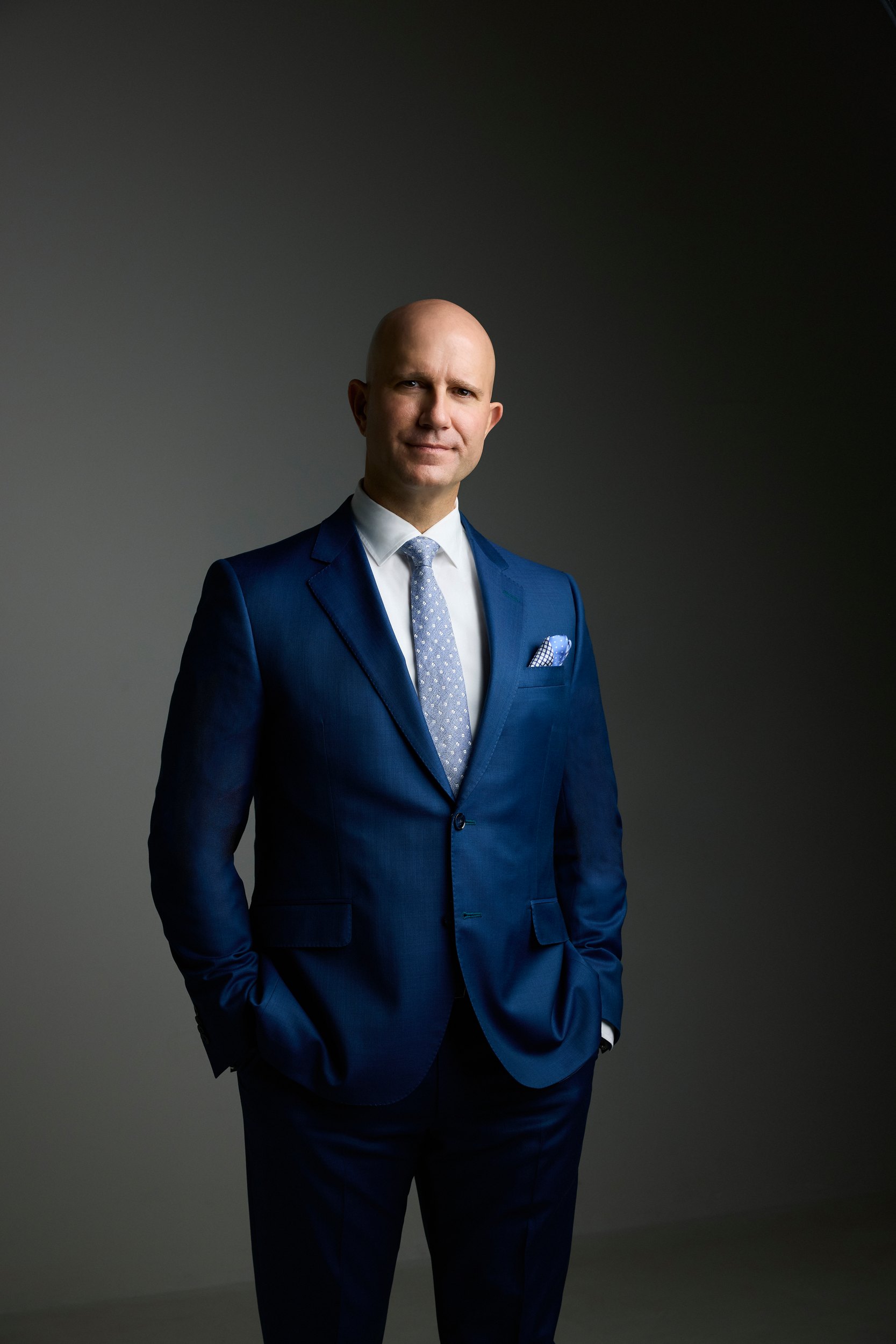Leadership portrait: standing, a navy suit on charcoal, hands in his pockets