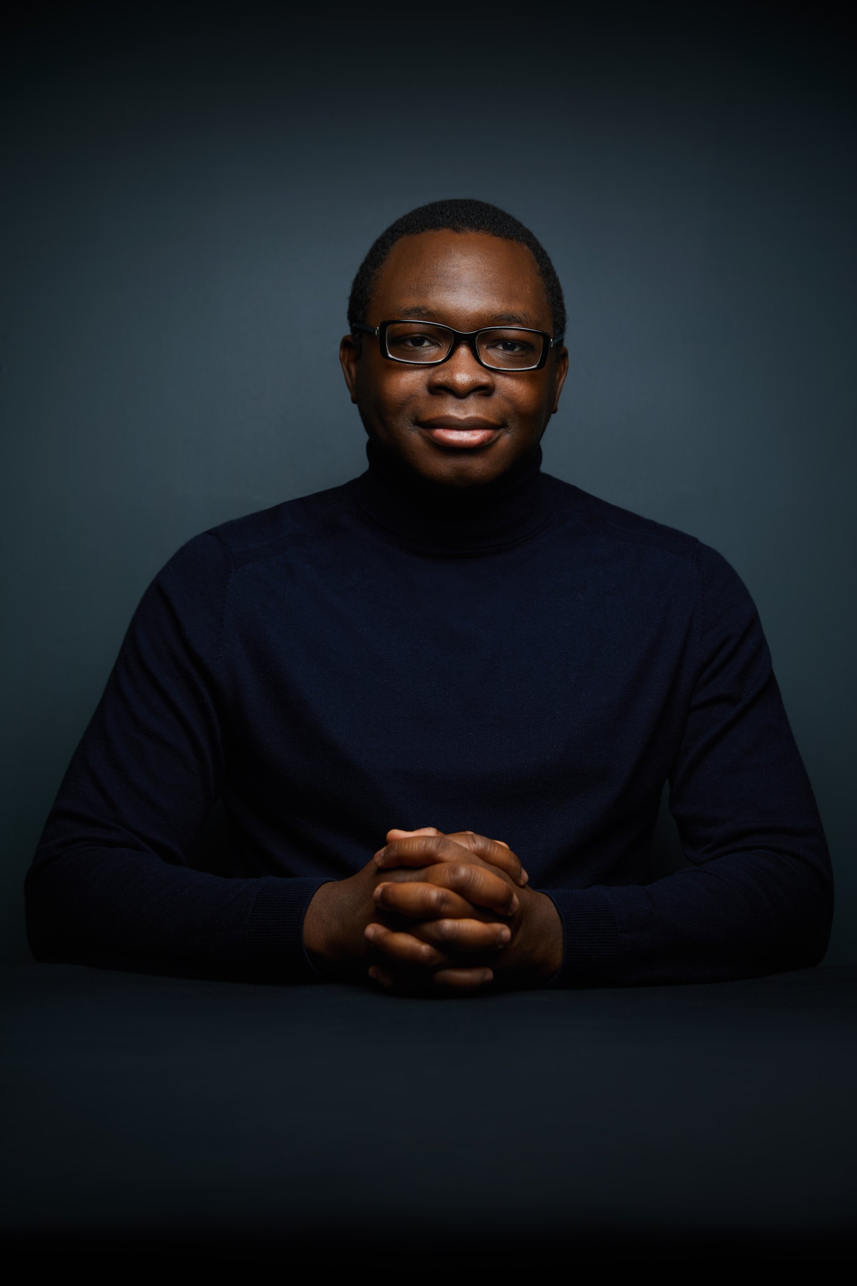 Leadership portrait: seated in a black turtleneck, hands clasped, in low light