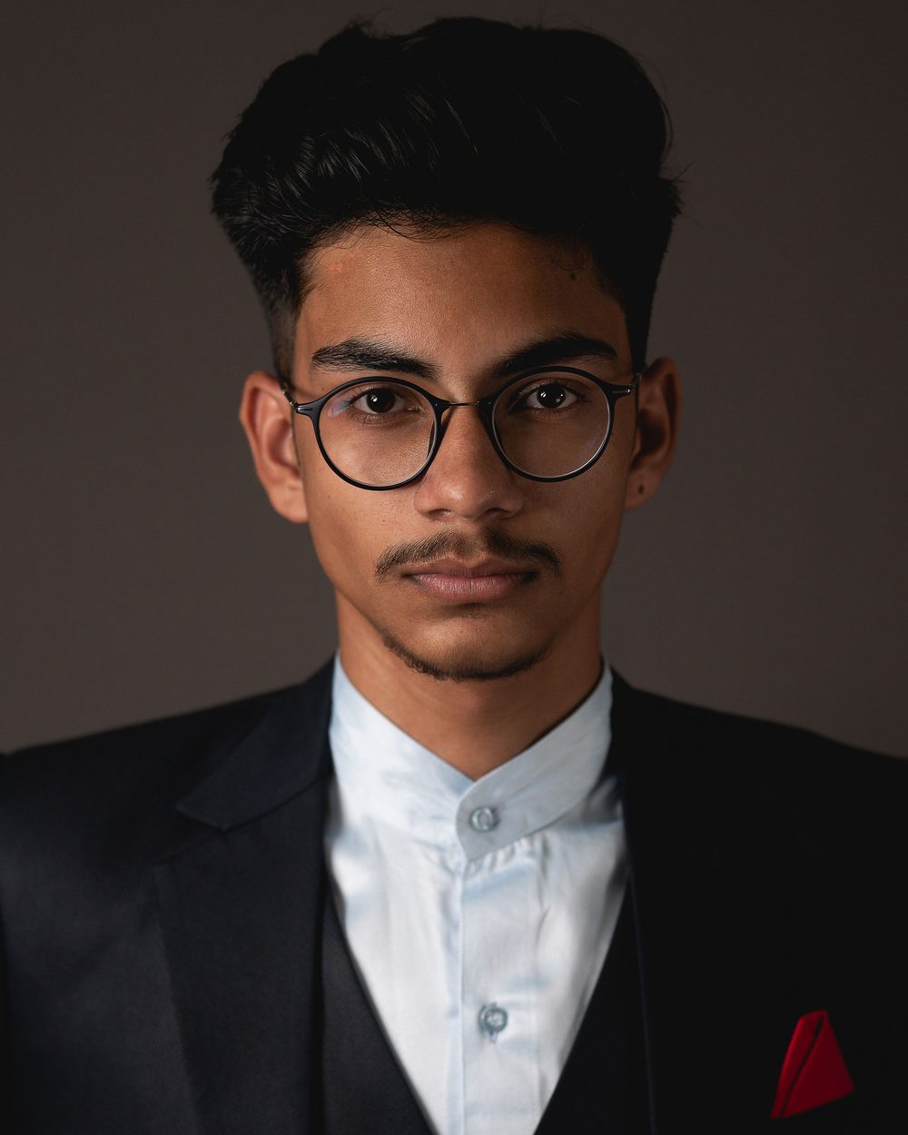Real photo: a young man with dark swept-back hair, round glasses and a moustache, in a black suit with a red pocket square, against a dark background