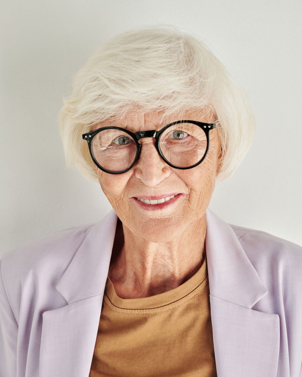 Real photo: a smiling older woman with short white hair and round black glasses, in a lilac blazer, against a white background