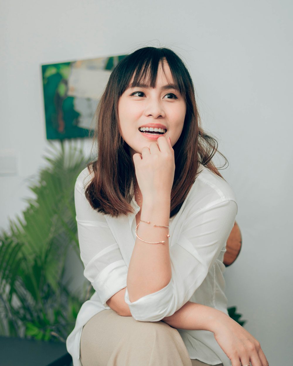 Real photo: a smiling woman with shoulder-length dark hair in a white blouse, her chin resting on her hand, with plants behind her