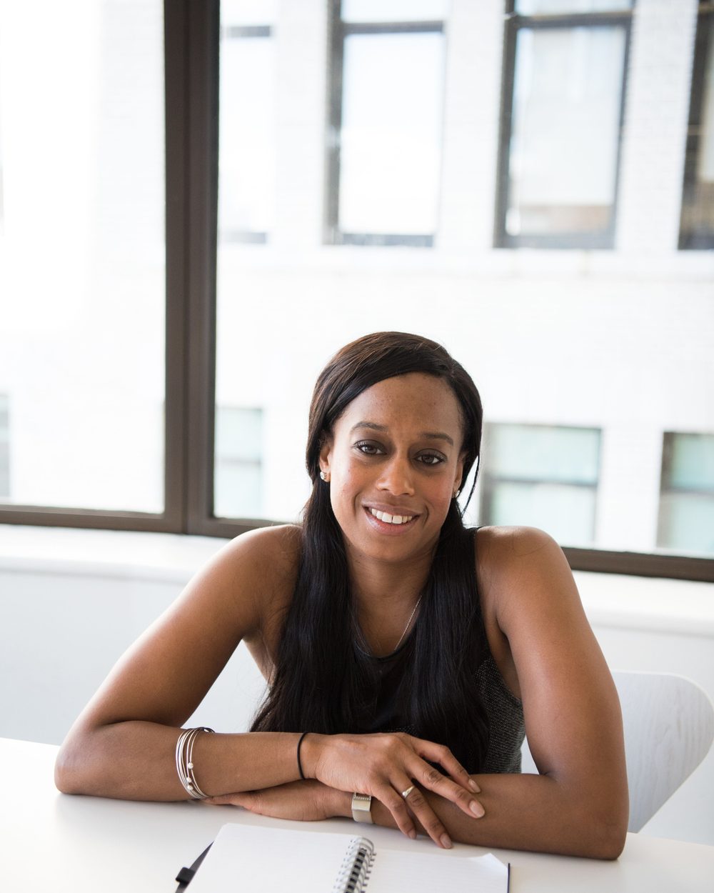 Real photo: a smiling woman with long dark hair leaning on a white desk by bright office windows