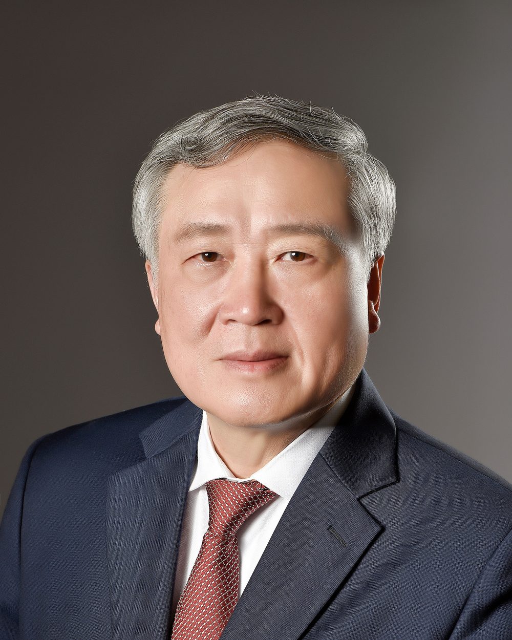 Real photo: an older man with grey hair in a navy suit, white shirt and red tie, against a grey background, his skin smoothed almost flat