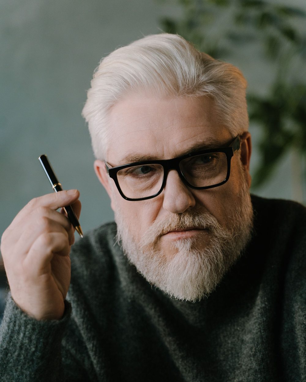 Real photo: an older man with white hair and a white beard, black glasses and a dark green sweater, holding a pen