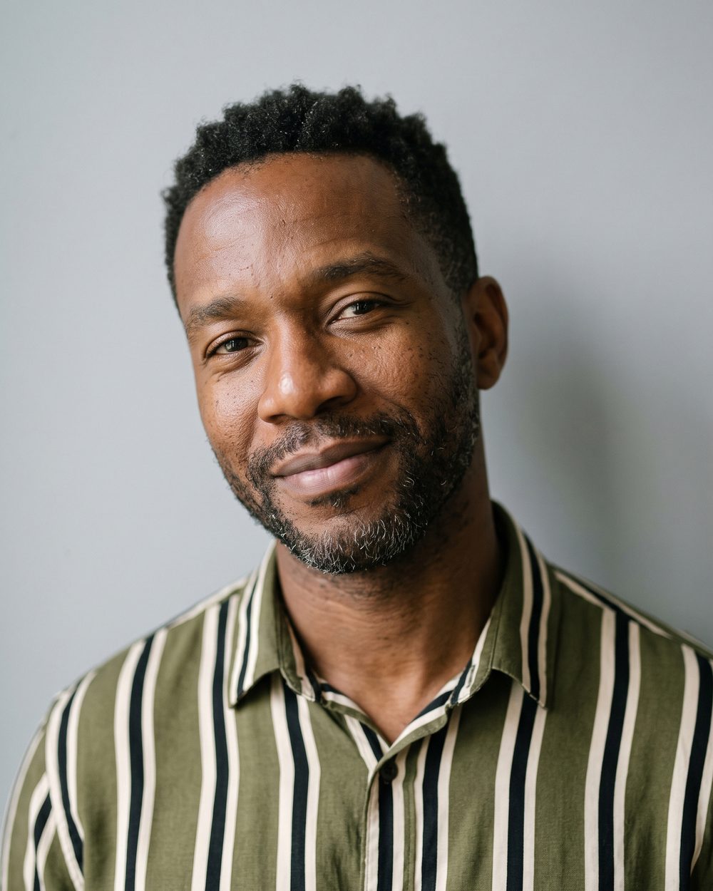 AI-generated face: a smiling man with a beard in a striped shirt, against a pale wall