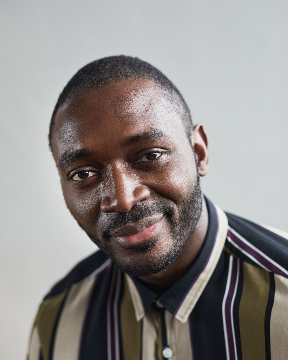 Real photo: a smiling man with a beard in a striped shirt, against a pale wall