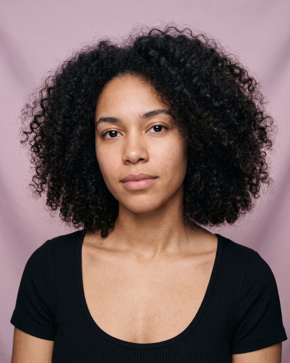 AI-generated face: a woman with dark curly hair and a black scoop-neck top, against a pink backdrop