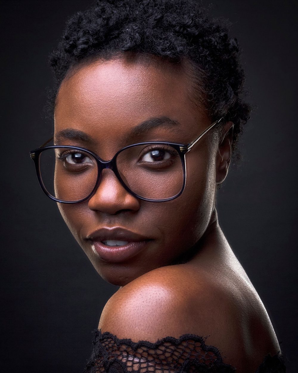Real photo: a woman with short natural hair and large black glasses looking over a bare shoulder, against a dark backdrop