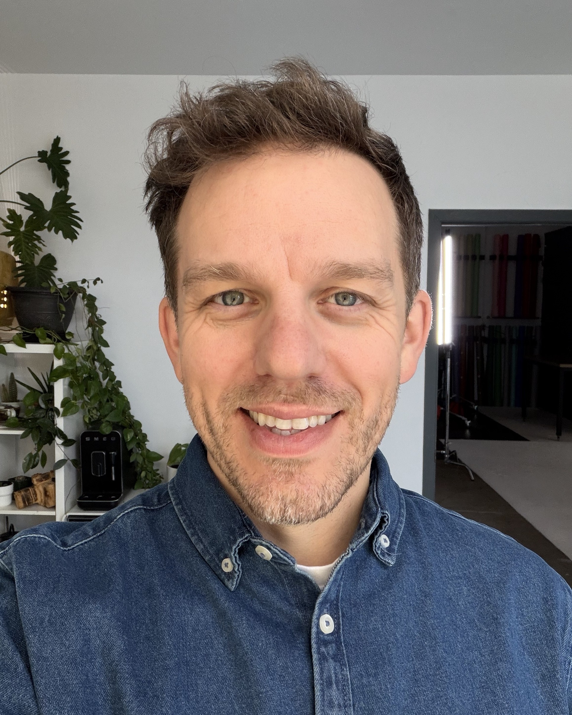 Real photo: Ryan Parker’s phone selfie in his studio, smiling, in a denim shirt, with plants and a coffee machine behind him
