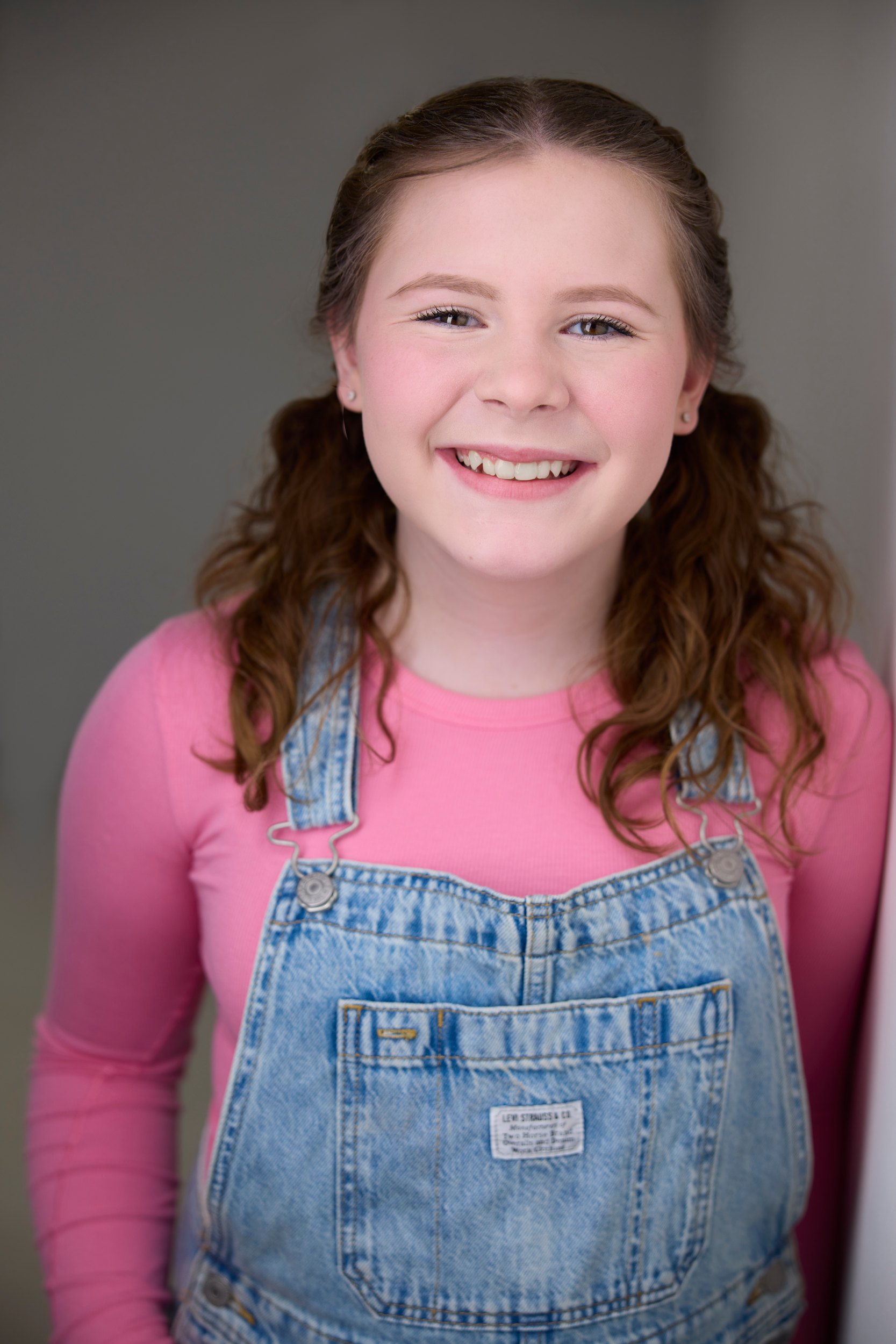 Kids and teens headshot: a pink top and overalls against the wall of light, pigtails, grinning