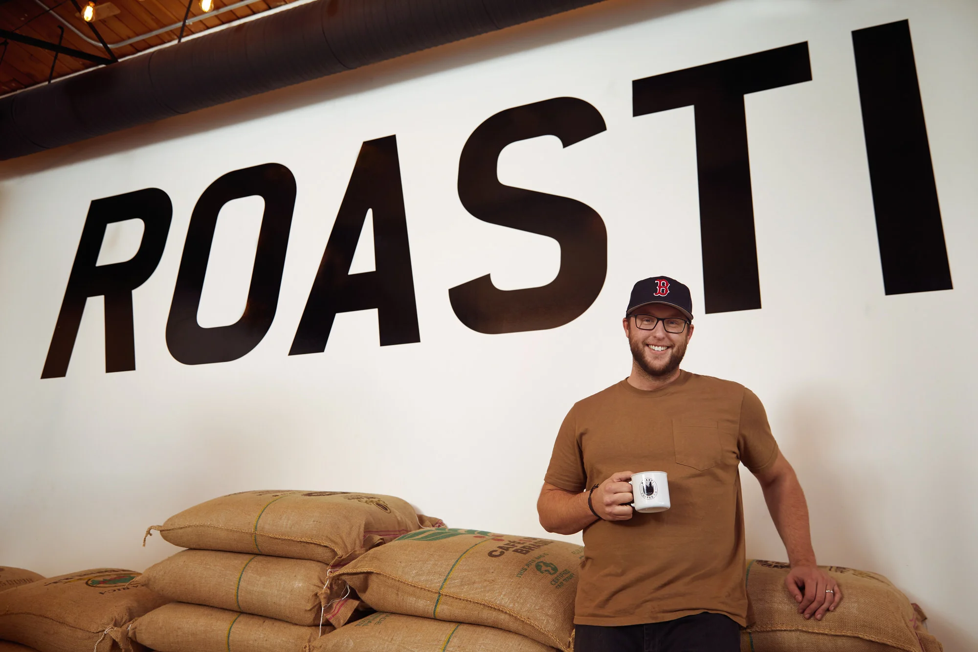 Under the Roasti wall sign, a mug in hand, among the coffee sacks