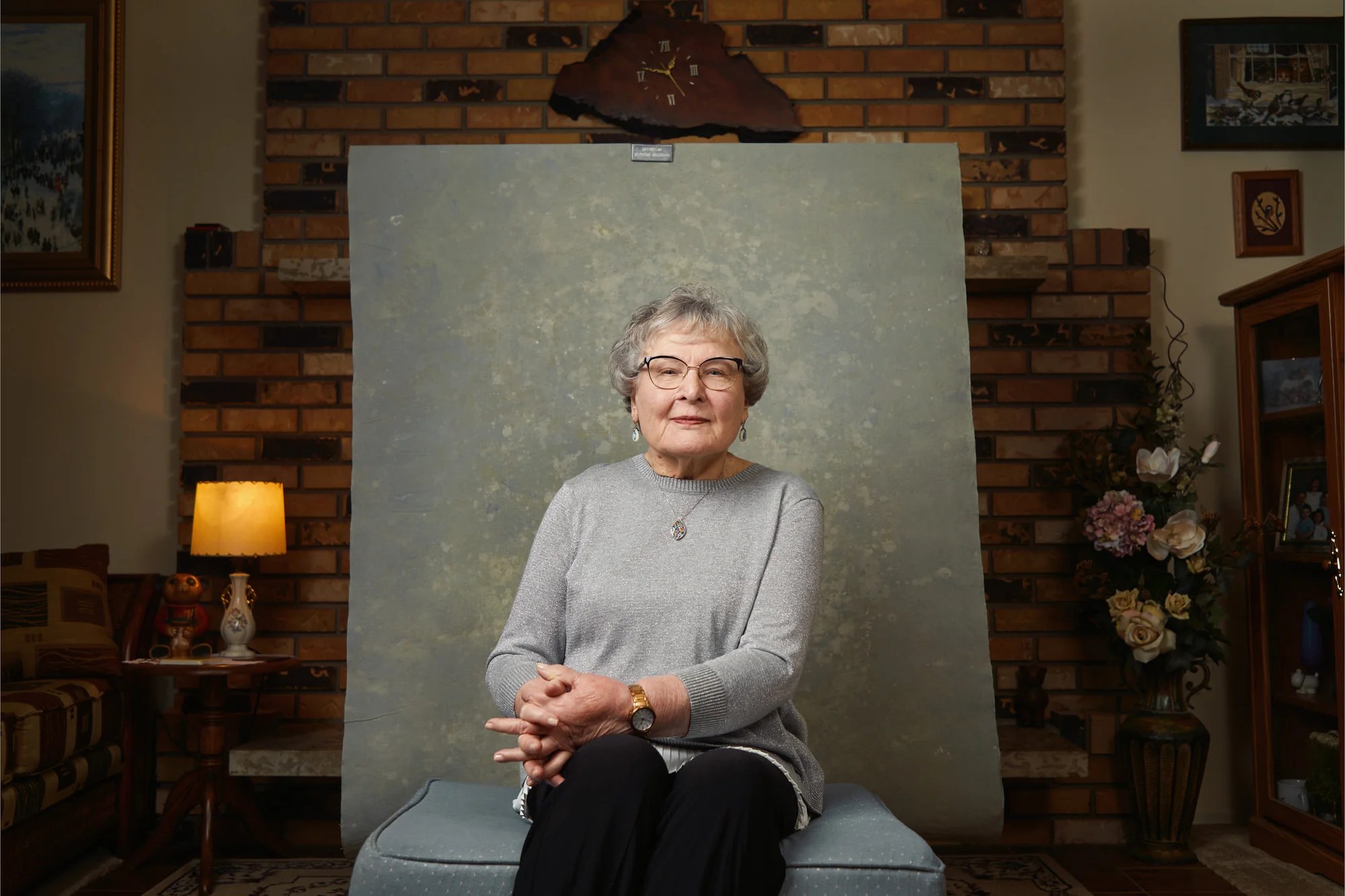 At home, seated in front of a hung backdrop, hands folded