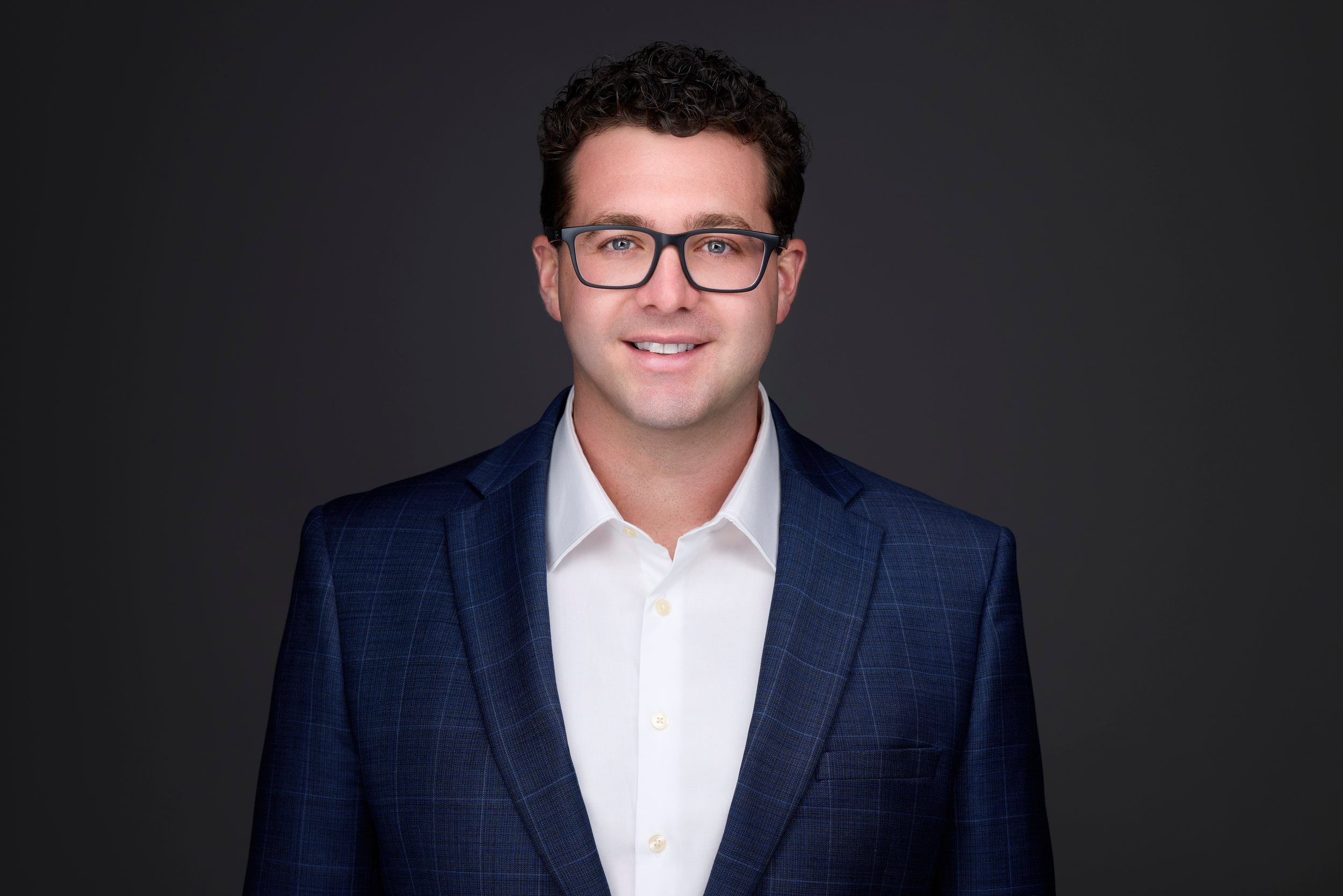 Real estate headshot: glasses, a navy check blazer and open white collar, on dark grey