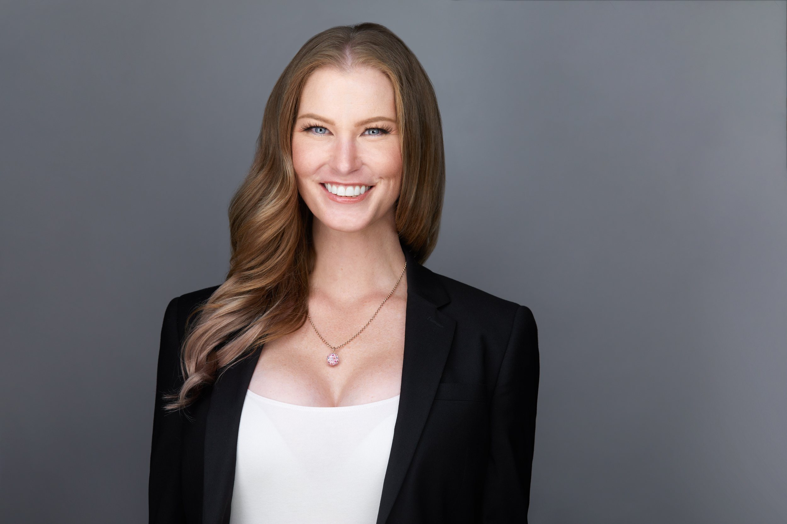 Real estate headshot: long hair, a black blazer over white, smiling, on grey
