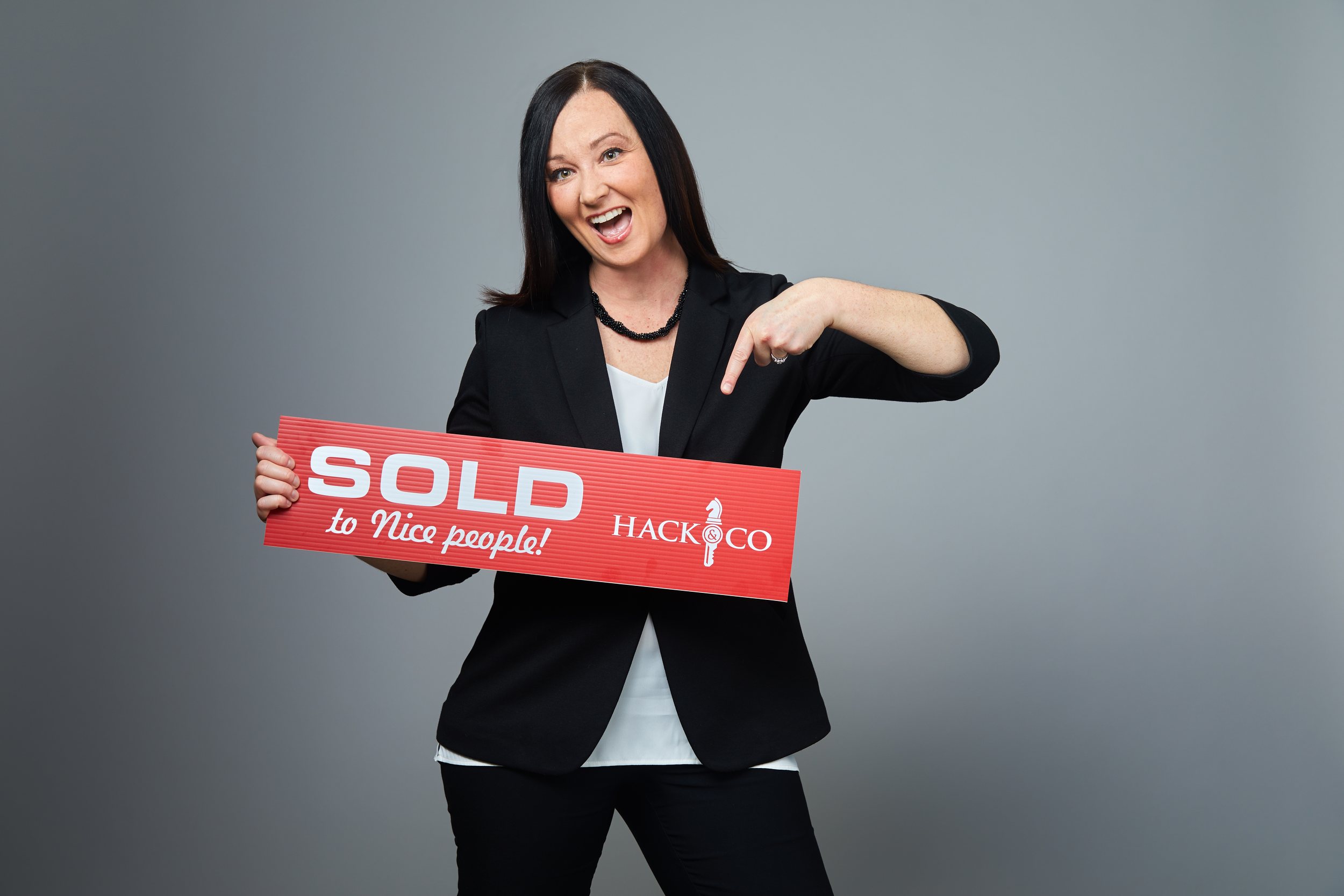 A real estate agent laughing and pointing at the SOLD sign she is holding, on grey