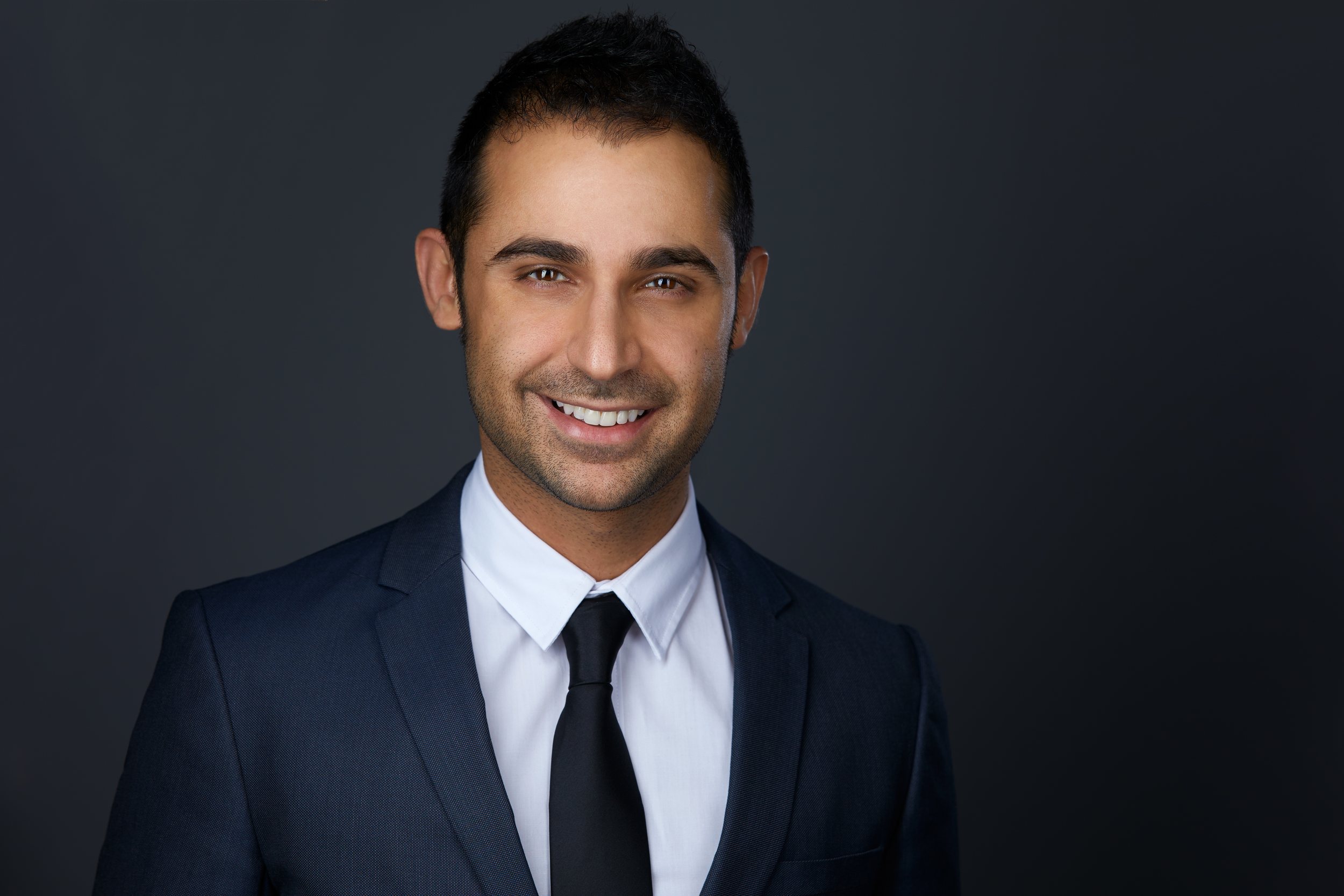 Real estate headshot: a dark suit and black tie, smiling, on dark grey