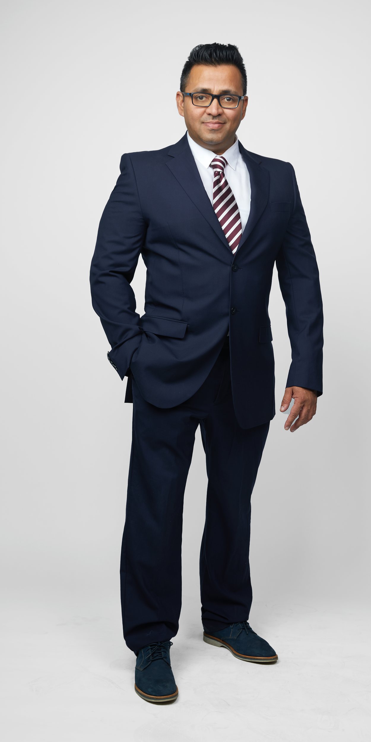 Full-length portrait on light grey: glasses, a navy suit and a striped tie, one hand in a pocket