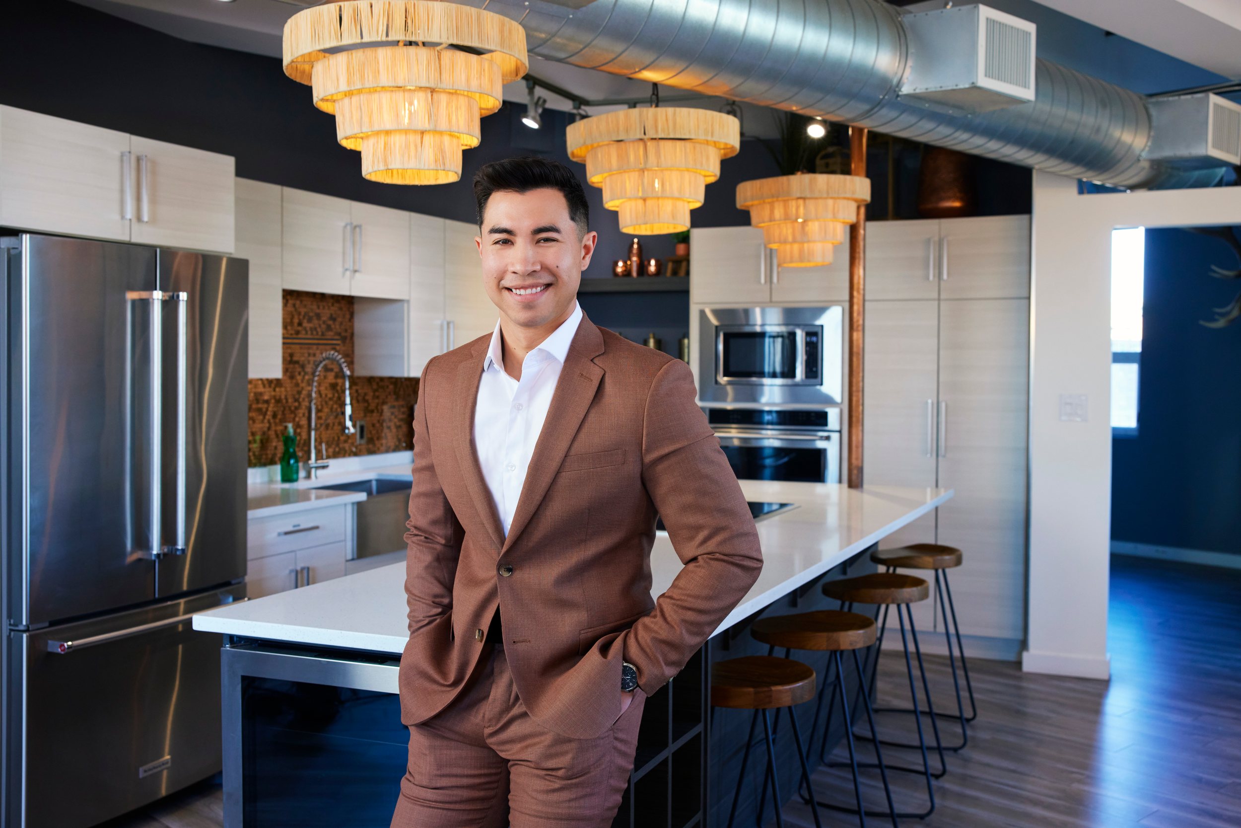 A realtor in a brown suit standing in a bright kitchen under gold pendant lights