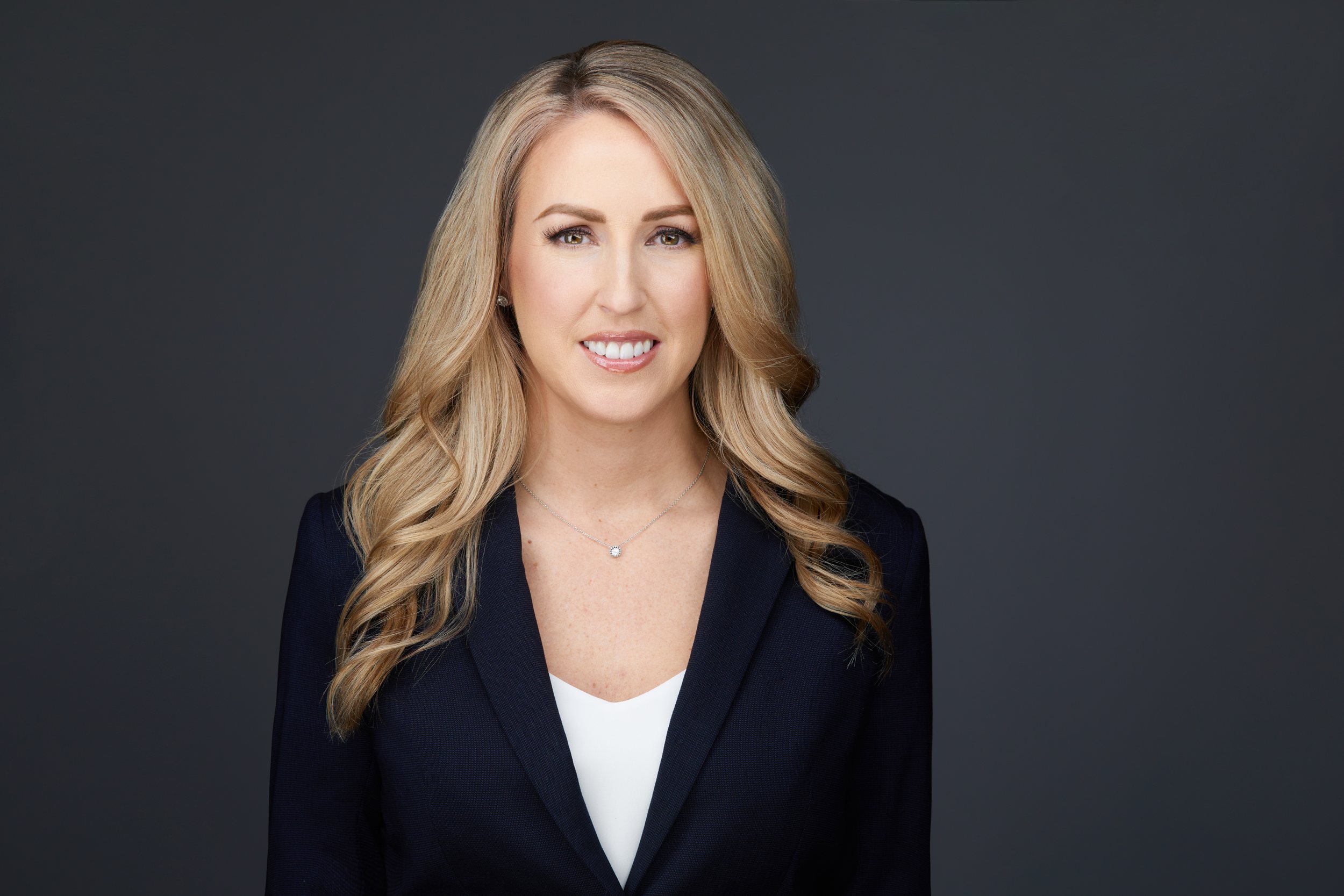 Real estate headshot: long blonde waves, a navy blazer over white, on dark grey