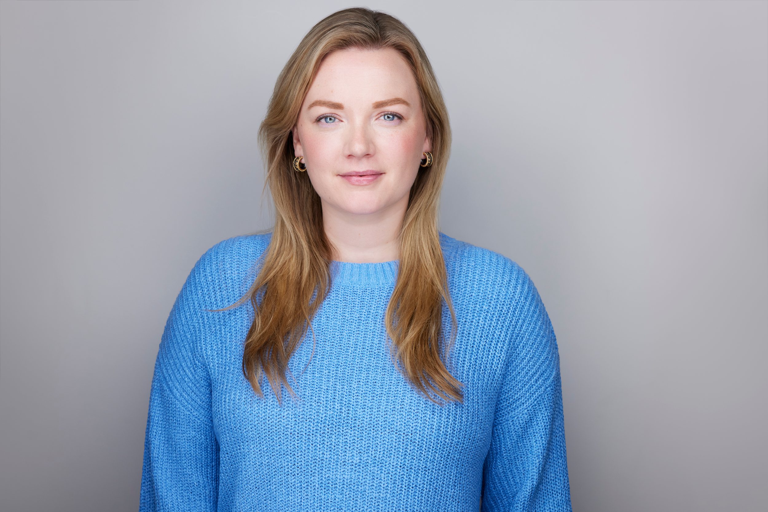 Real estate headshot: long strawberry blonde hair, a blue knit sweater, on light grey