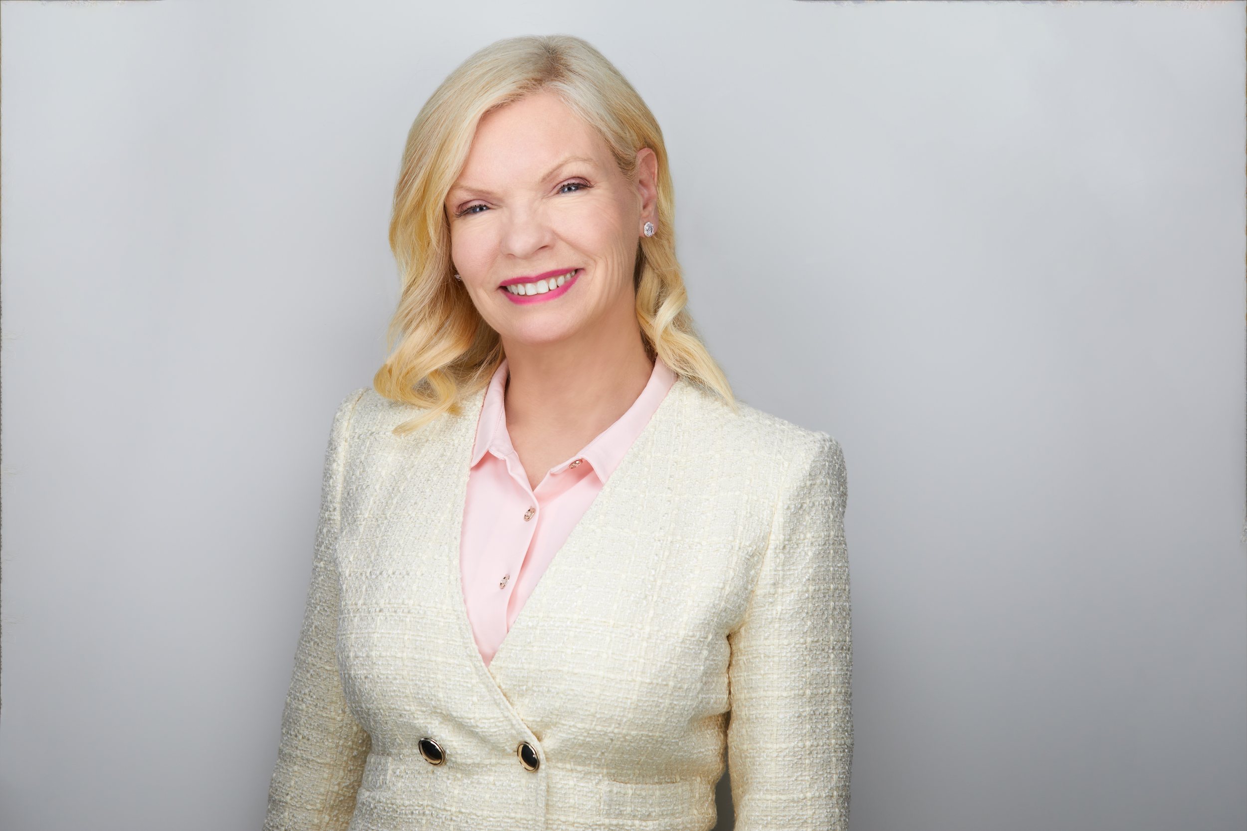 Real estate headshot: blonde hair, a cream tweed jacket over pink, on light grey