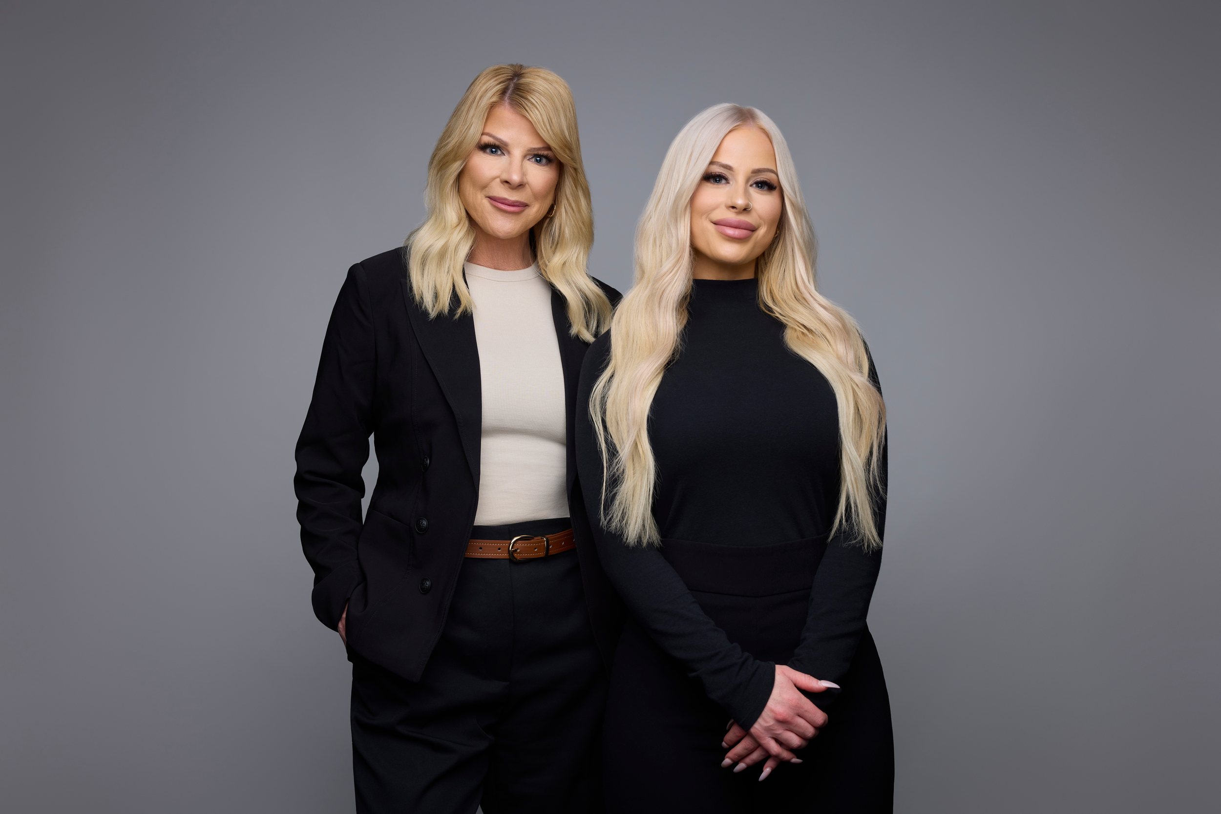 A mother and daughter real estate team side by side, long blonde hair and black blazers, on grey