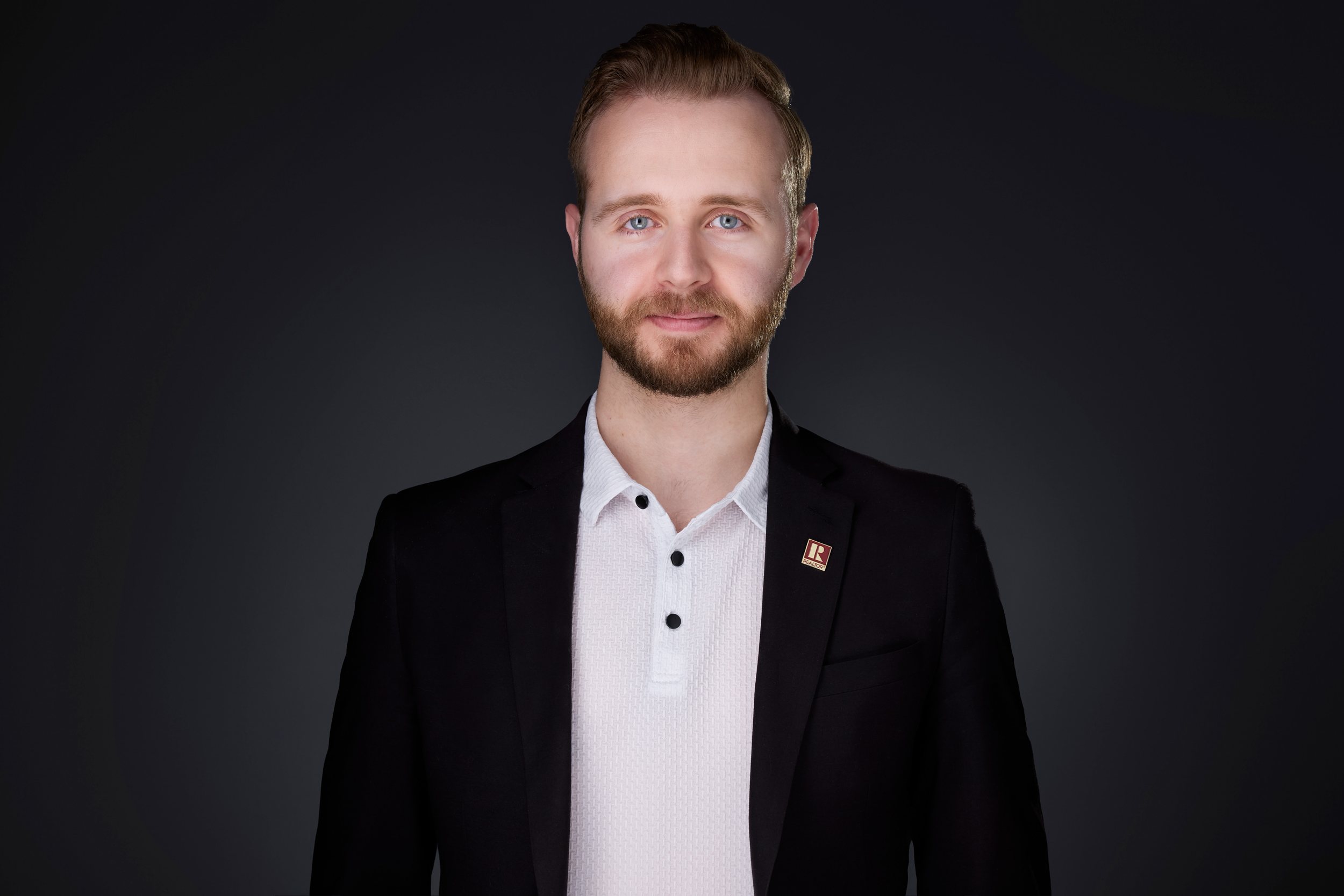 Real estate headshot: a beard, a black blazer with a lapel pin over a white collar, on dark