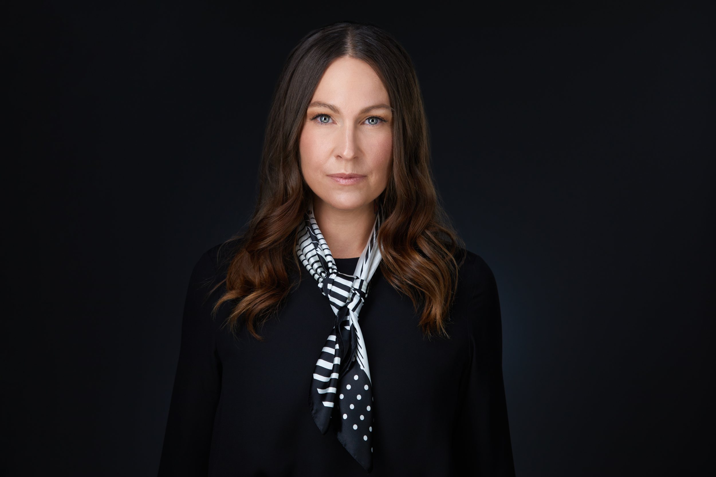 Real estate headshot: long brown hair, a patterned scarf and a black jacket, on dark