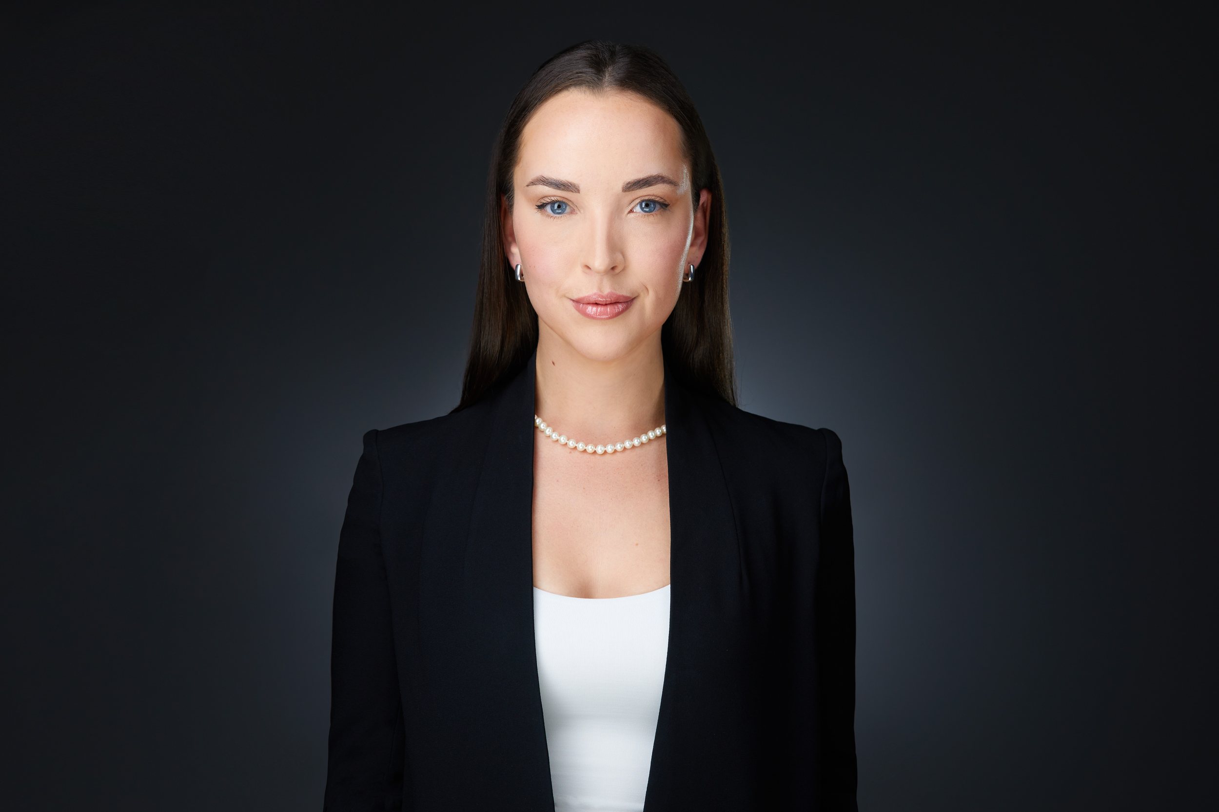 Real estate headshot: dark hair, a black blazer over white and a pearl necklace, on dark