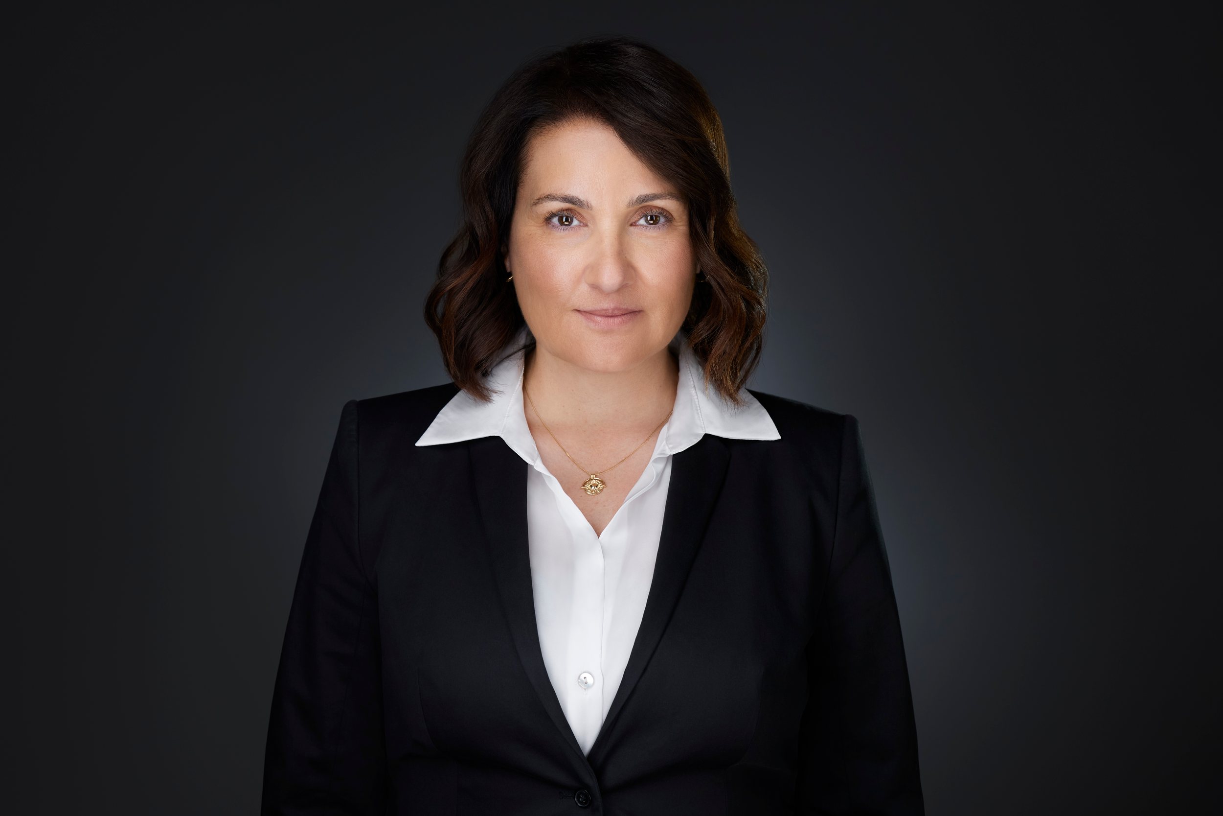 Real estate headshot: dark hair to the shoulders, a black blazer over a white blouse, on dark