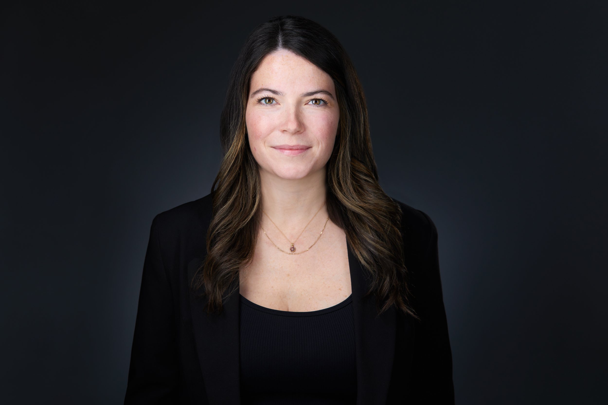 Real estate headshot: long dark hair, a black blazer and a pendant, on dark